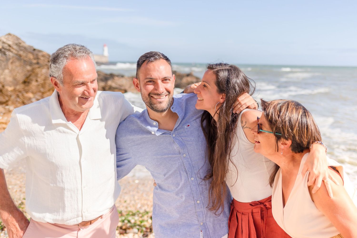 Four people share laughs on a sunny beach with waves crashing against rocks in the background.
