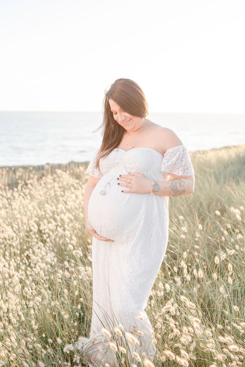 Glowing maternity photo of woman in lace dress against ocean backdrop.
