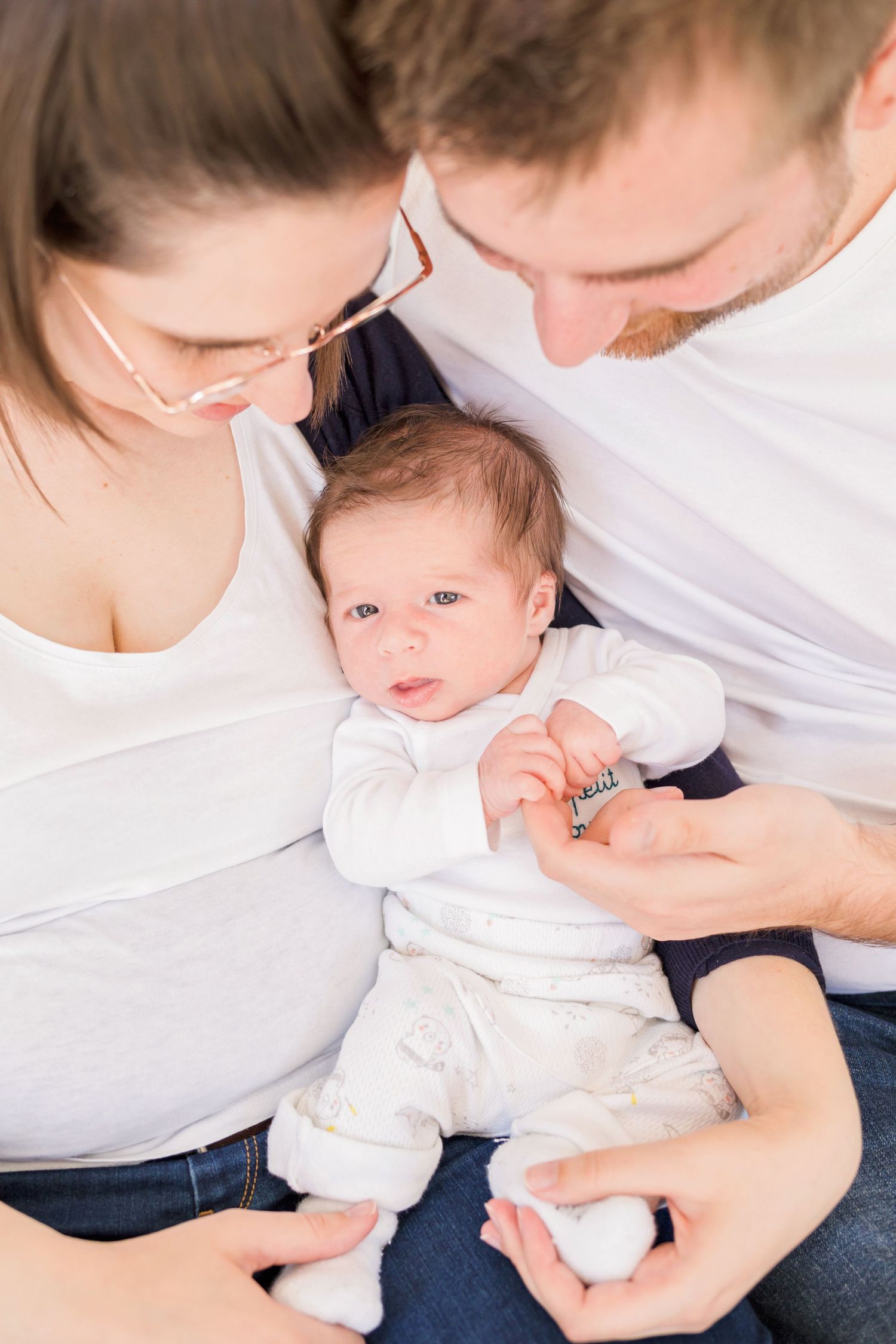 Parent in white shirt and jeans holding small baby in white clothing while sitting on floor.