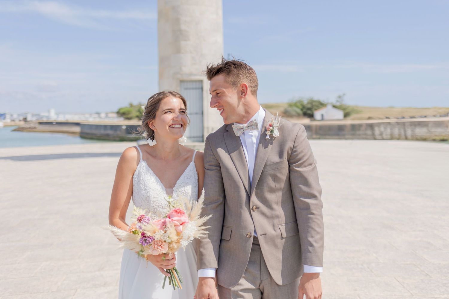 Newlyweds share joyful moment together near lighthouse by harbor.
