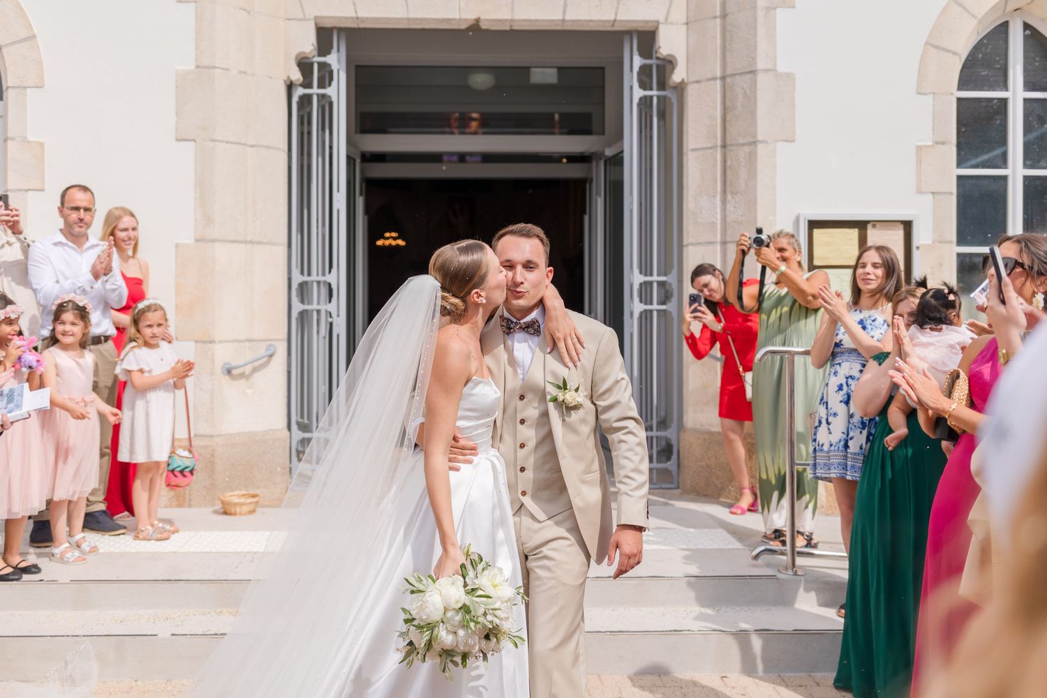 A wedding couple shares a joyful embrace outside a stone church building while guests look on.