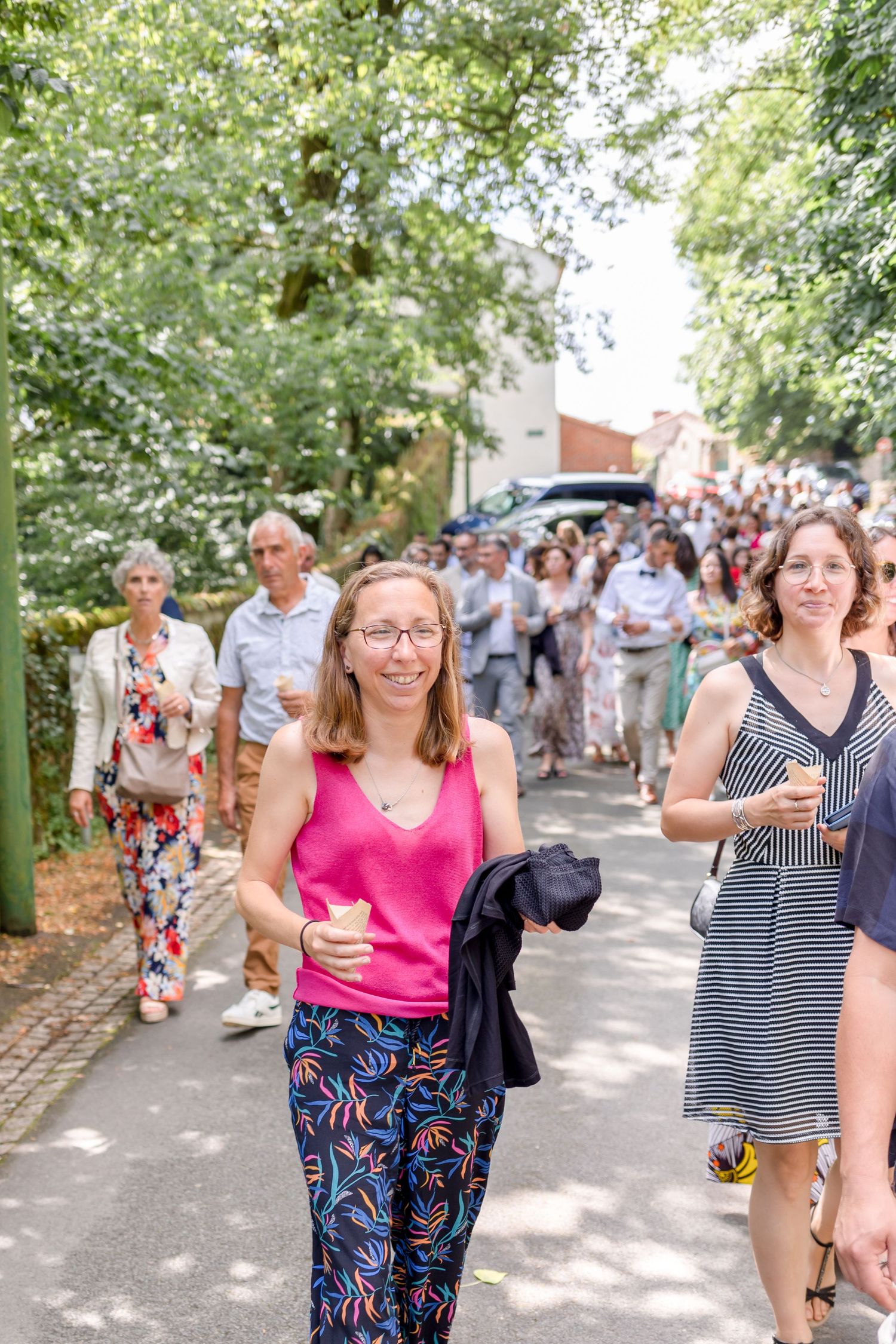 Wedding guests walking along sunny tree-lined path during celebration.