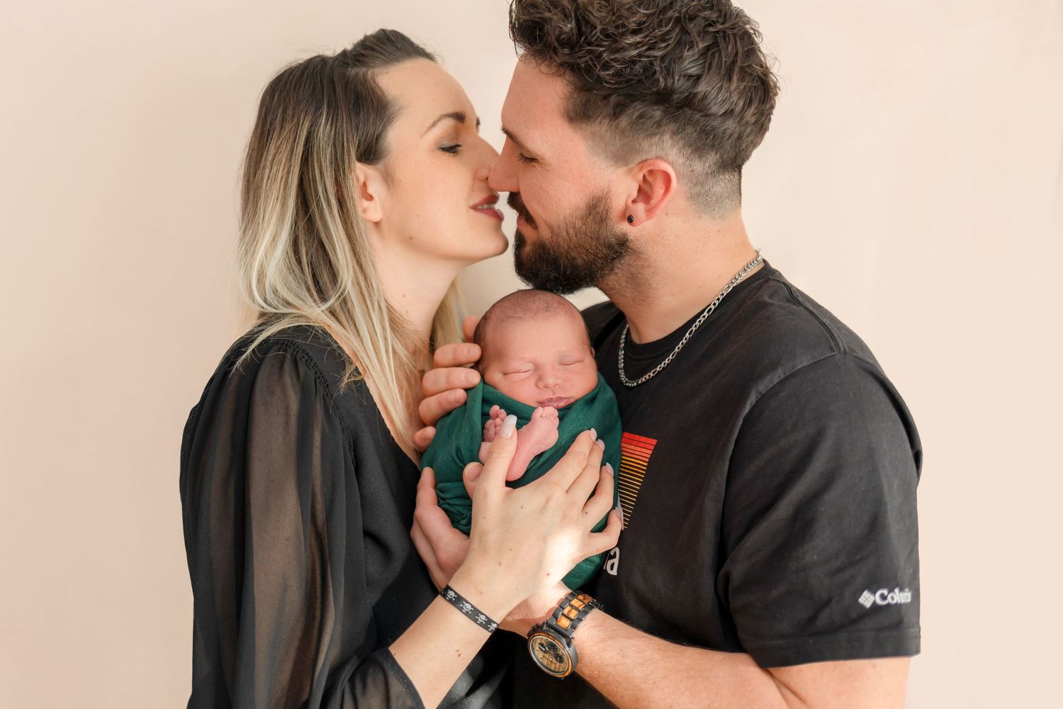 Parents share an affectionate moment while holding their sleeping baby in green.