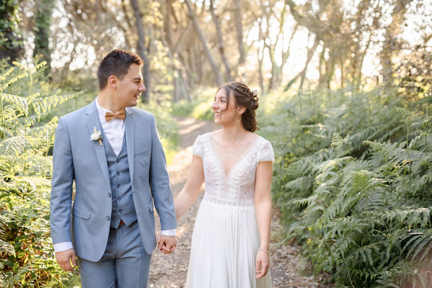 A wedding couple walks hand in hand through a forest path lined with ferns.