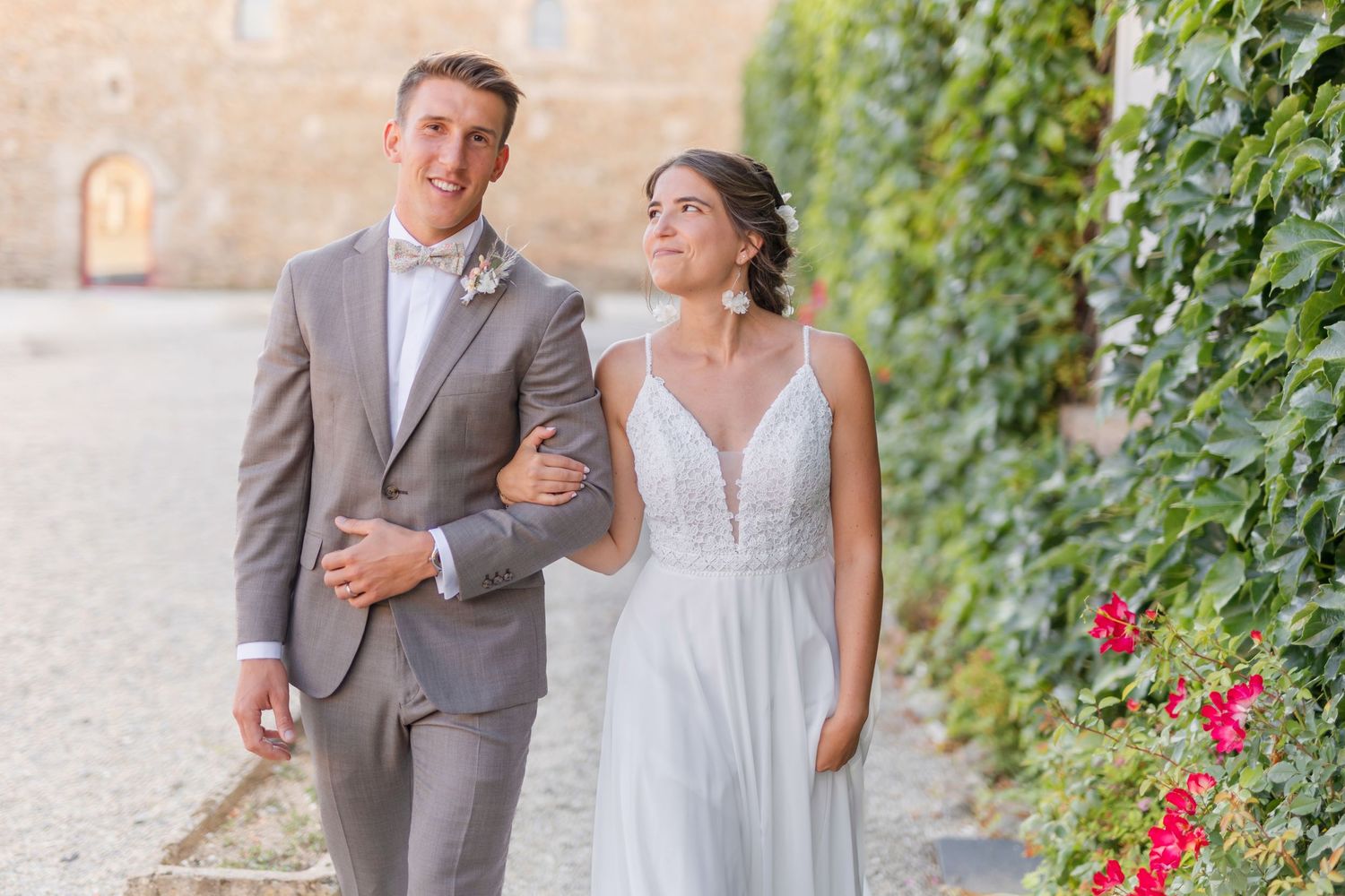 Wedding couple walking arm in arm along a vine-covered wall with red flowers.