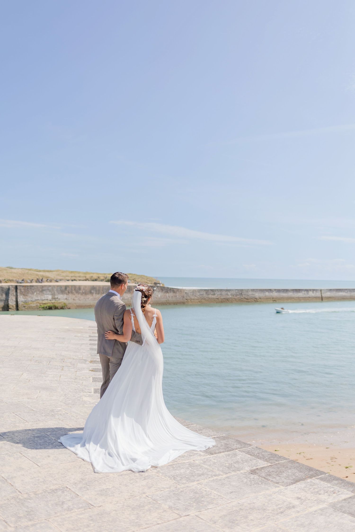 A wedding couple poses together on a sunny beach near the water's edge.