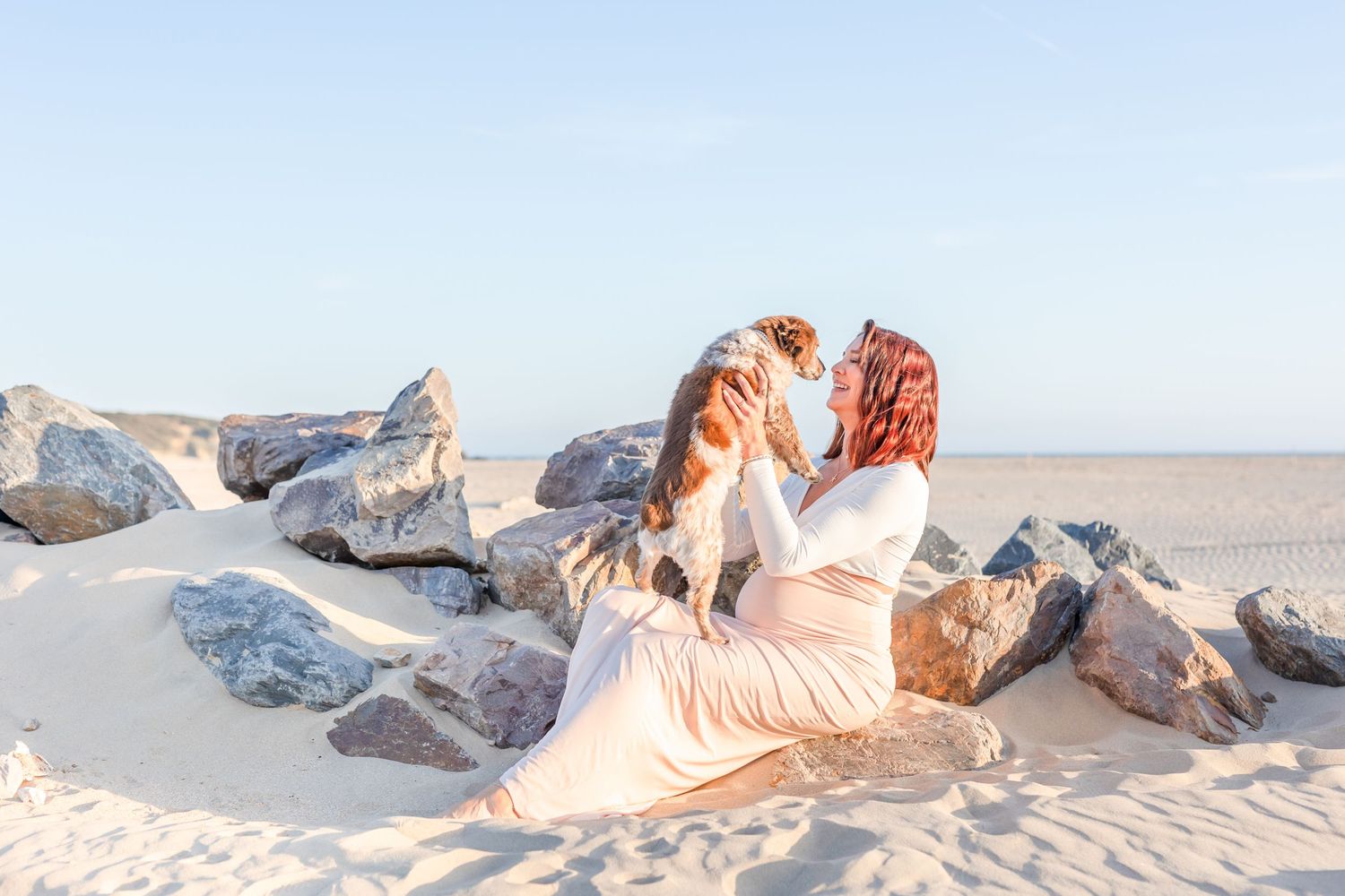 Woman in flowing dress sitting on coastal rocks holding a dog at sunset.
