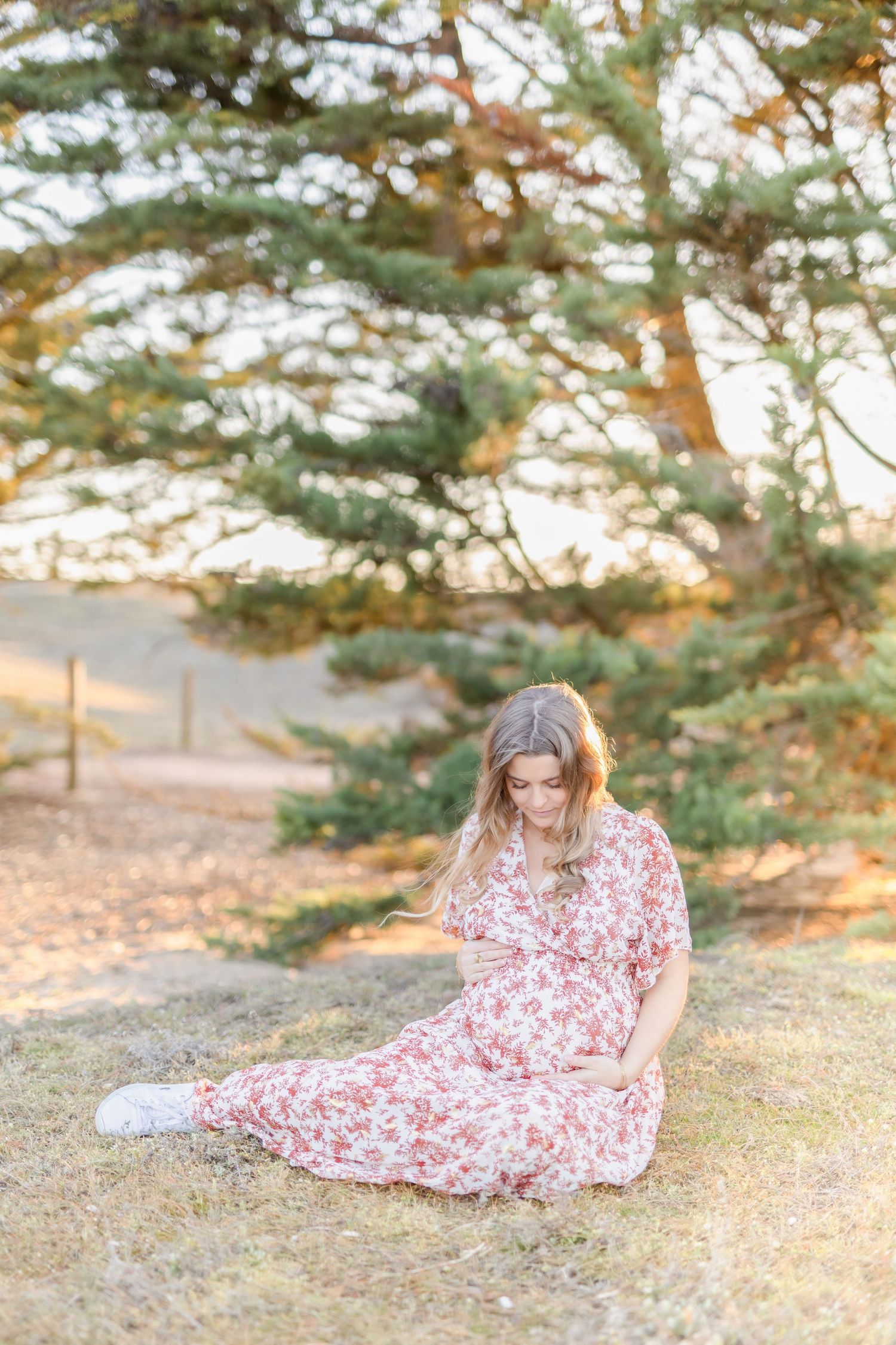 Woman in floral dress sitting peacefully on ground with pine trees in background.