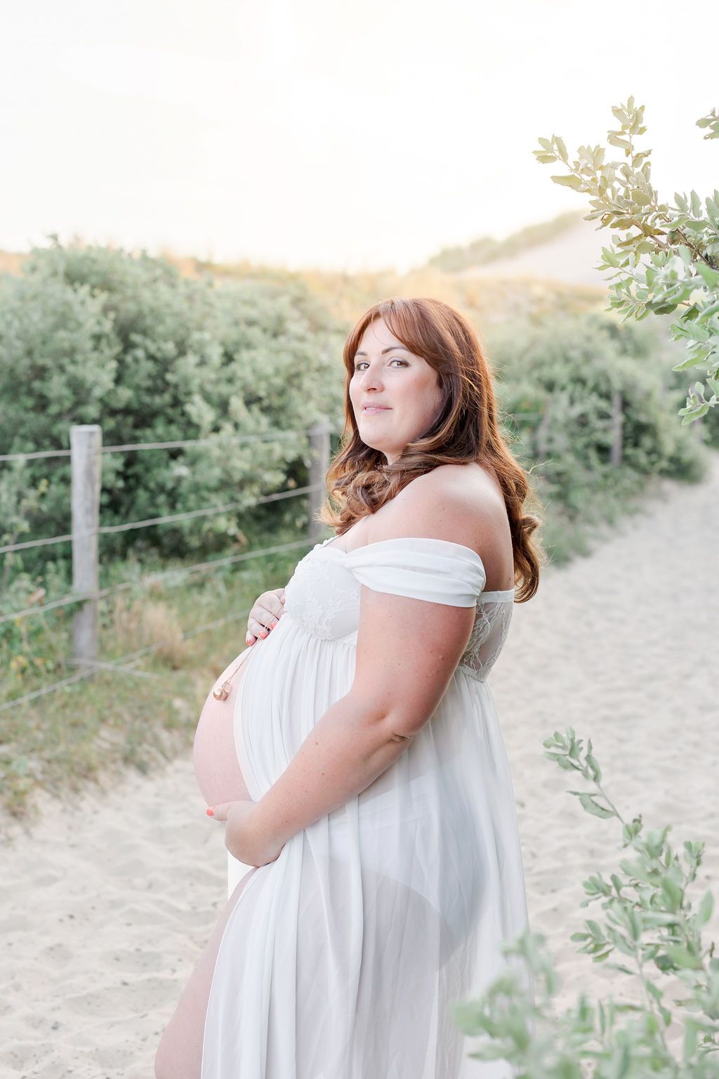 A serene maternity photo of a woman in white dress cradling her baby bump on a scenic trail.