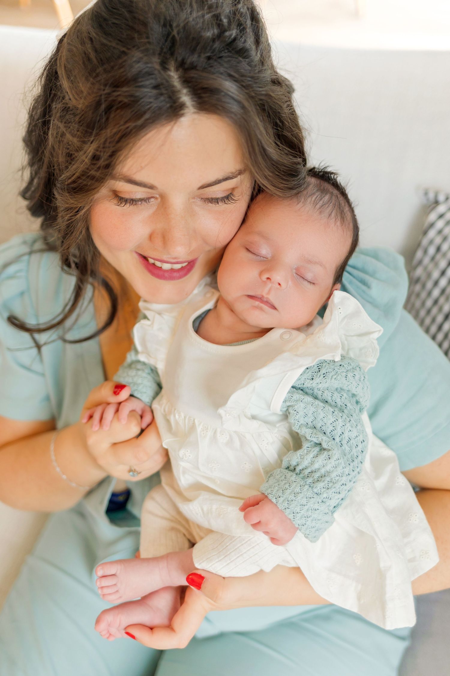 Mother in mint blue dress holding sleeping newborn in white lace outfit.