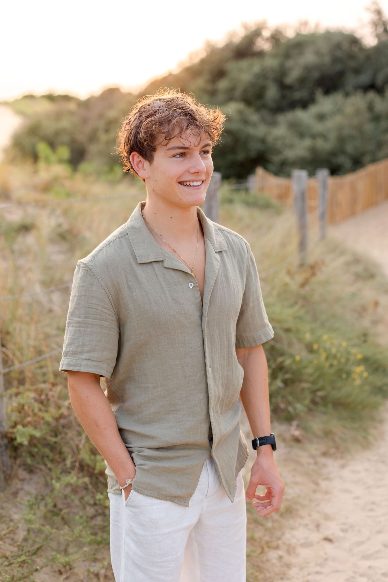 Person in casual beige shirt smiling while standing on dirt path with greenery in background.
