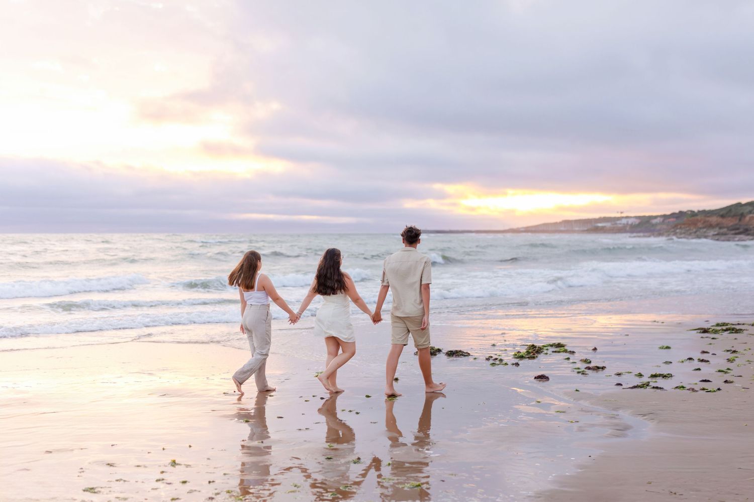 Three people holding hands while walking on beach during golden hour sunset.