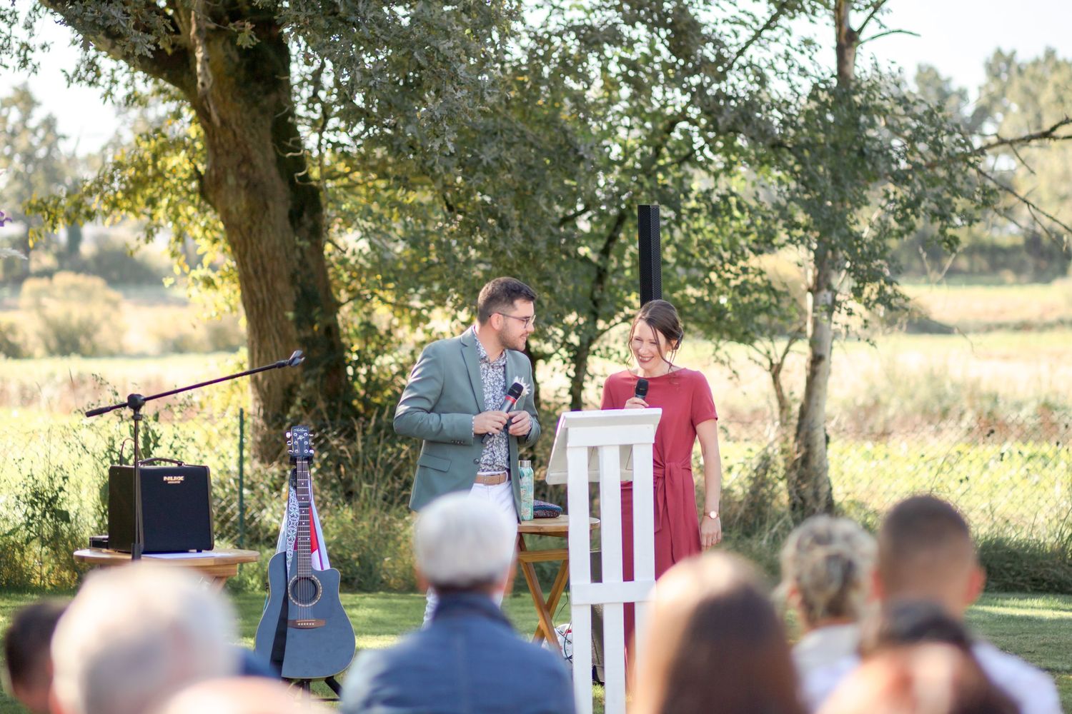 Speakers address wedding guests at outdoor ceremony podium with guitar visible.