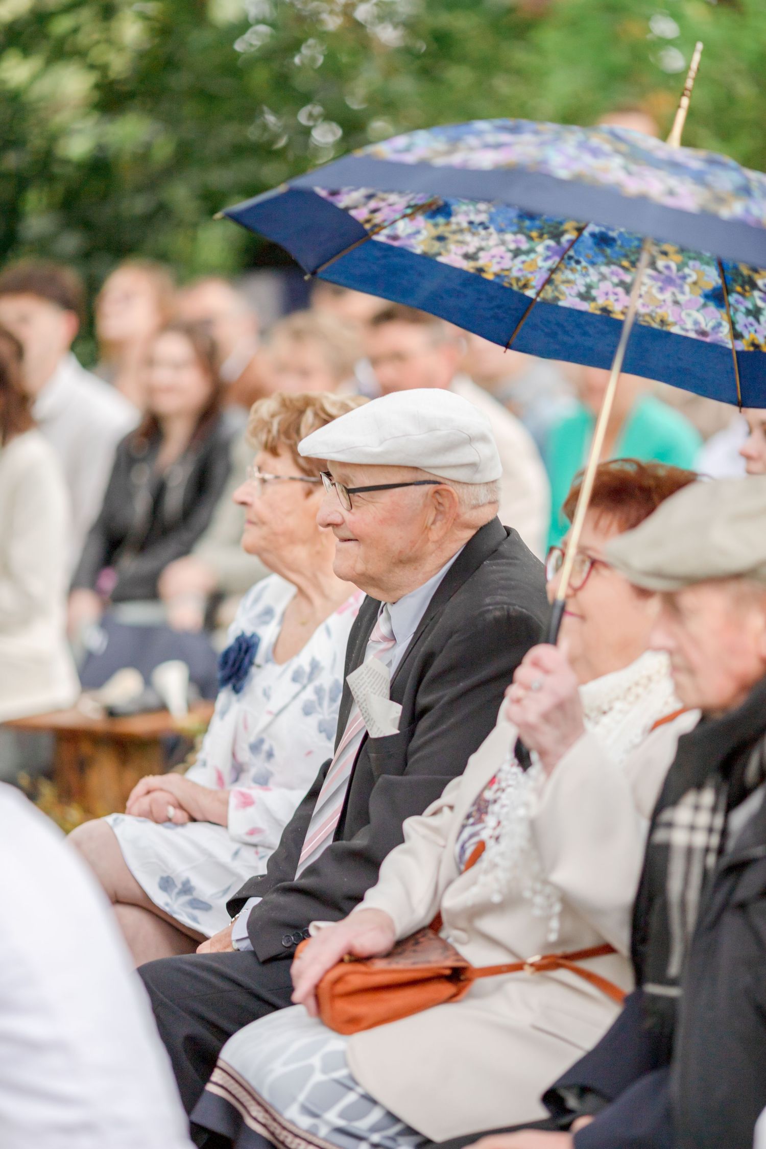 Guests sit under umbrellas at an outdoor event in formal attire.
