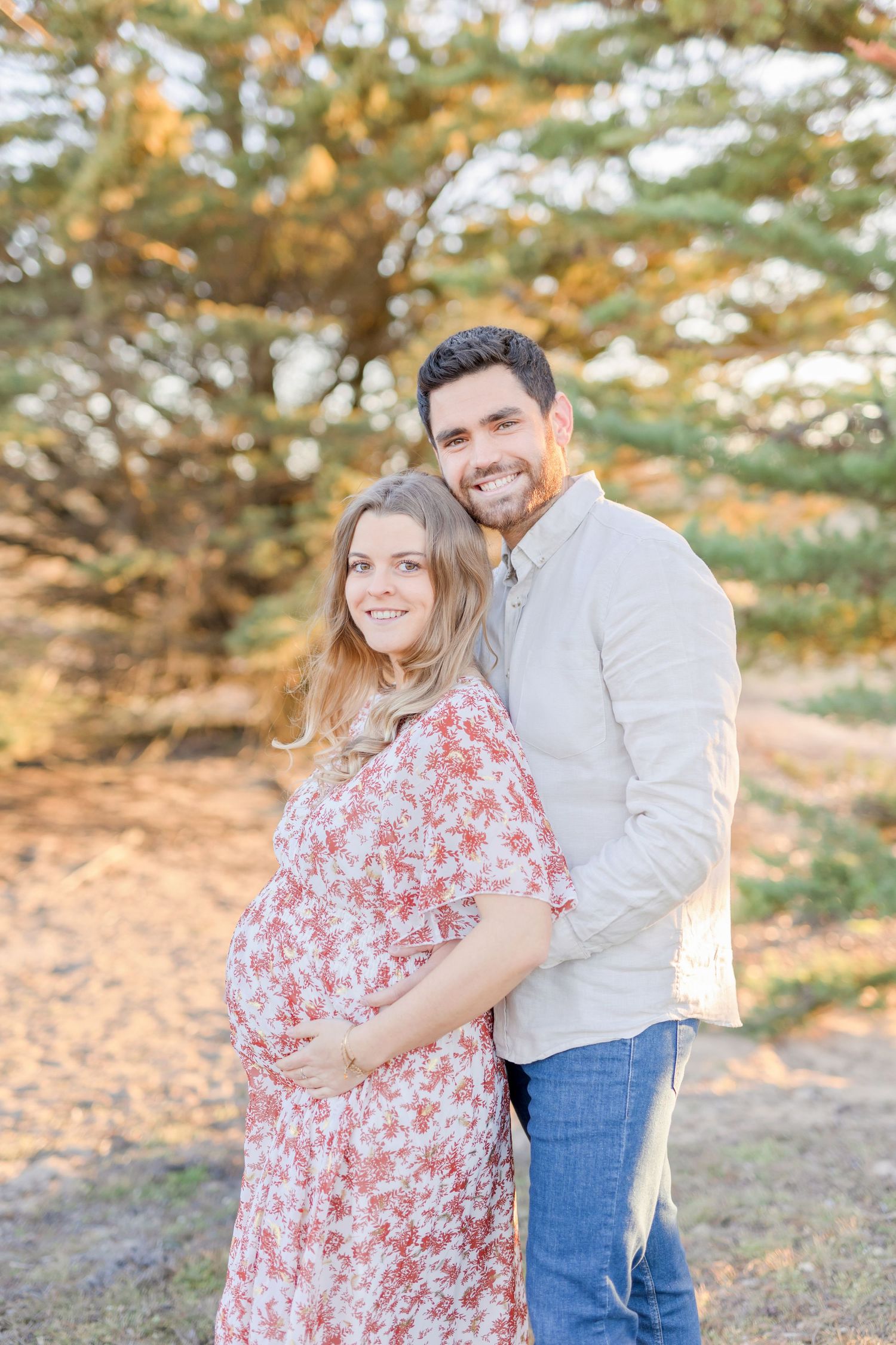 Expecting couple poses for photo in nature with warm sunset lighting.