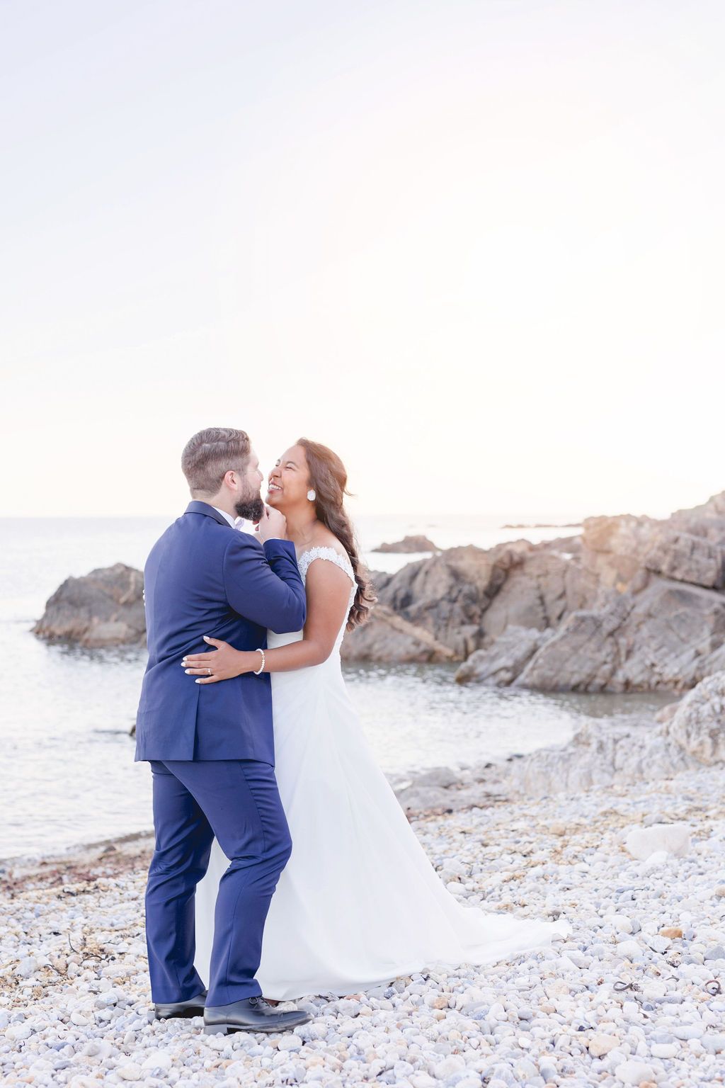 A seaside wedding portrait with couple embracing on pebbled beach at sunset.