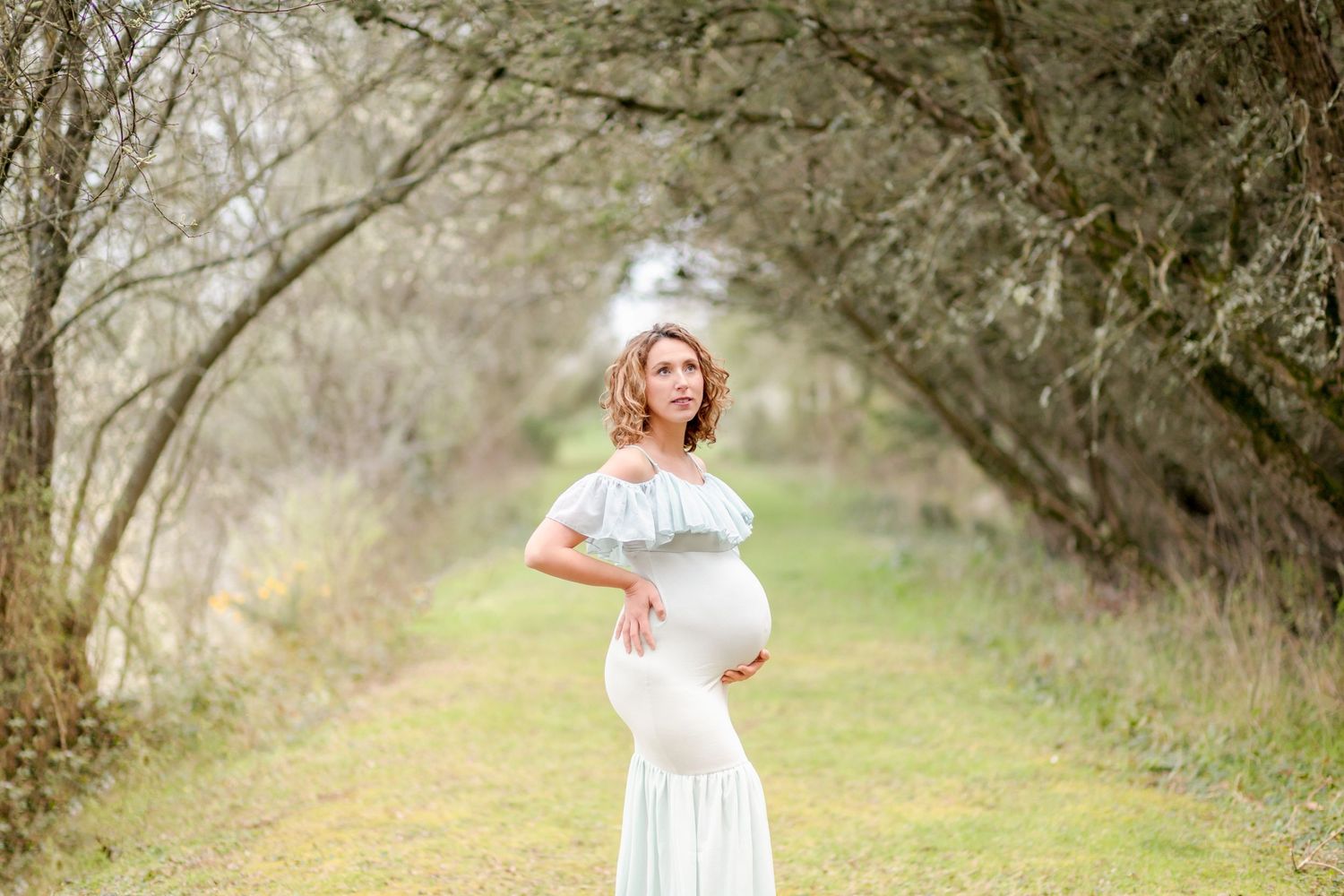 Ethereal maternity photo under an archway of trees in soft natural lighting.