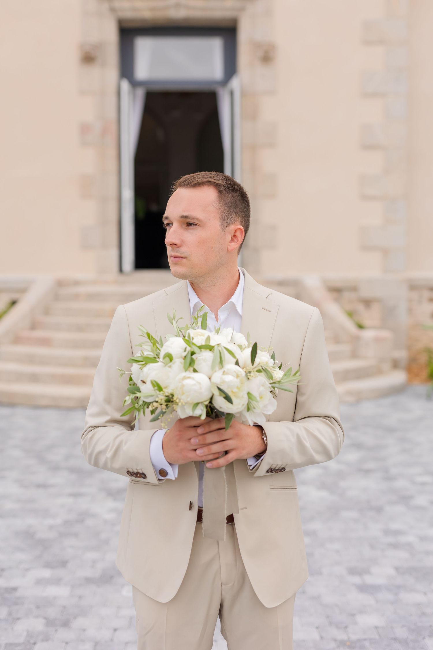 Person in beige suit holding white floral bouquet outside stone building.
