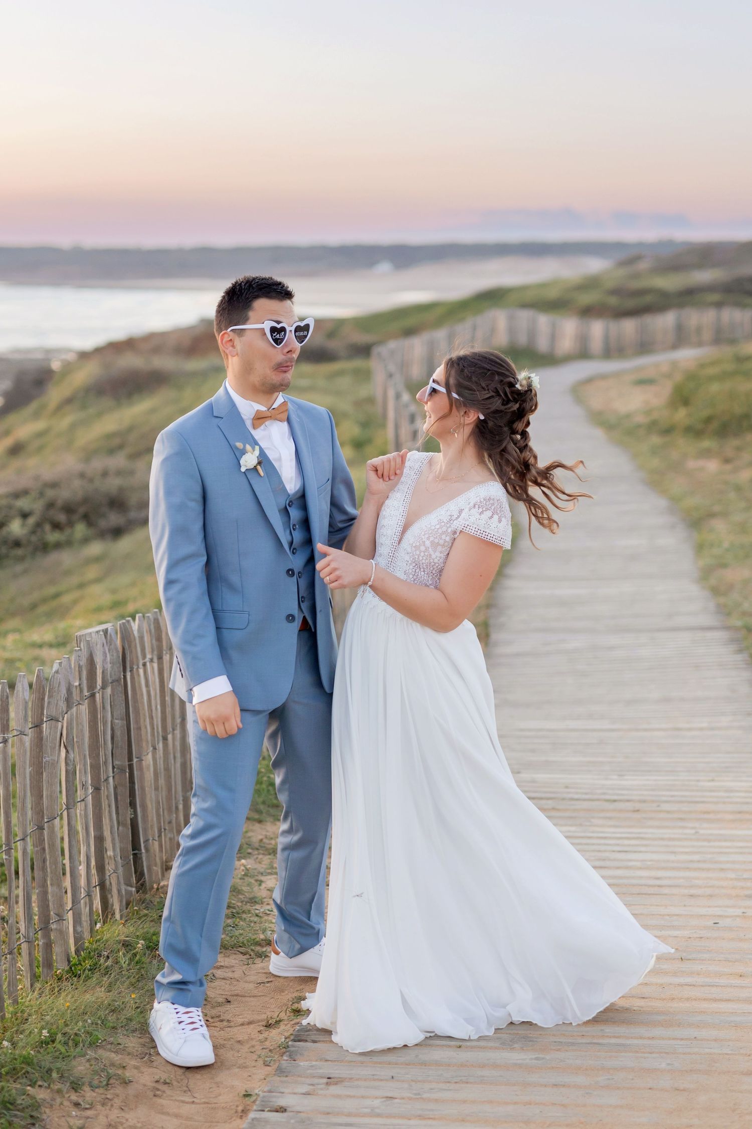 Playful wedding couple wearing sunglasses on seaside boardwalk at sunset.
