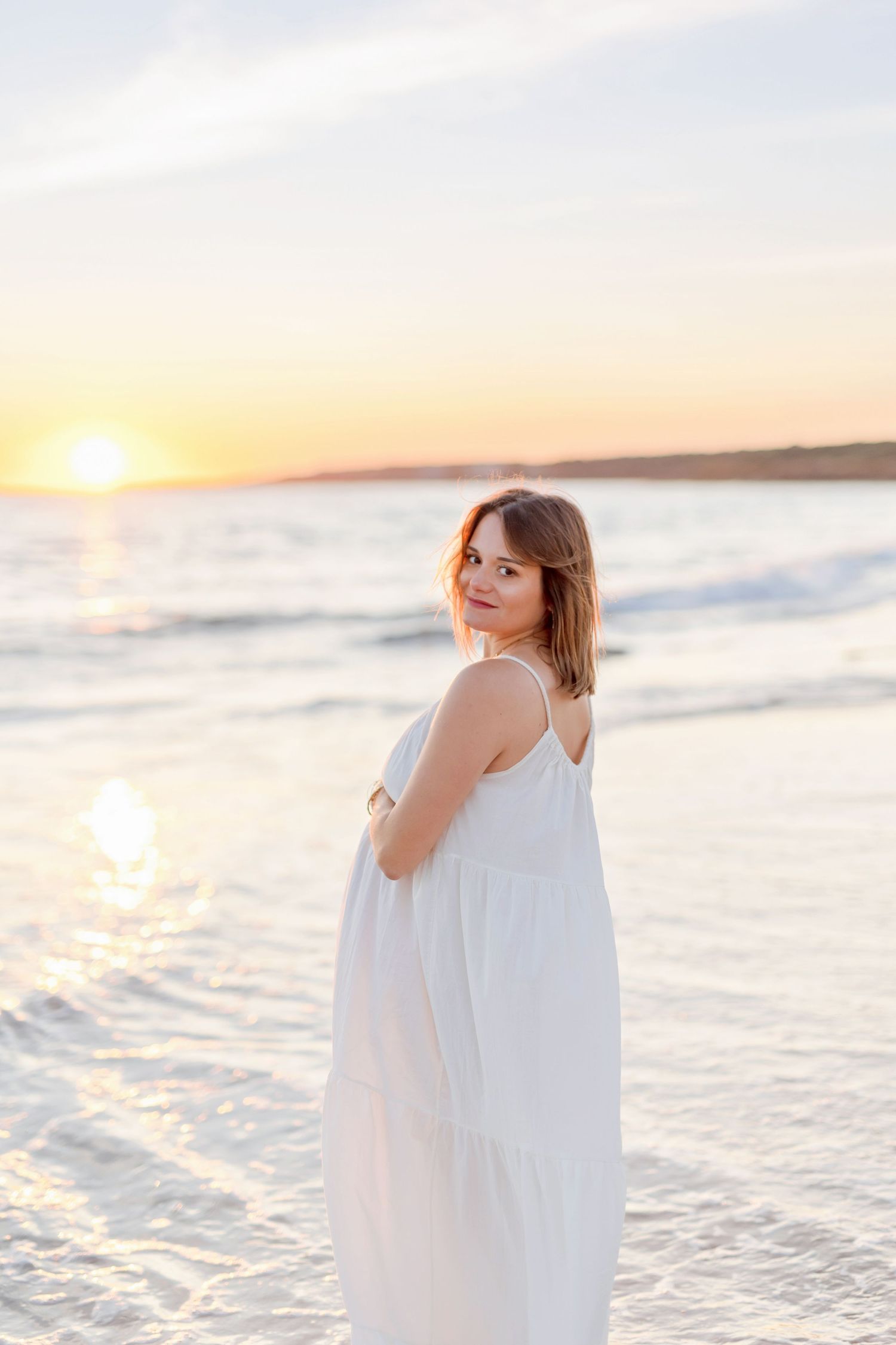 Woman in flowing white dress poses on beach at sunset with ocean waves behind.