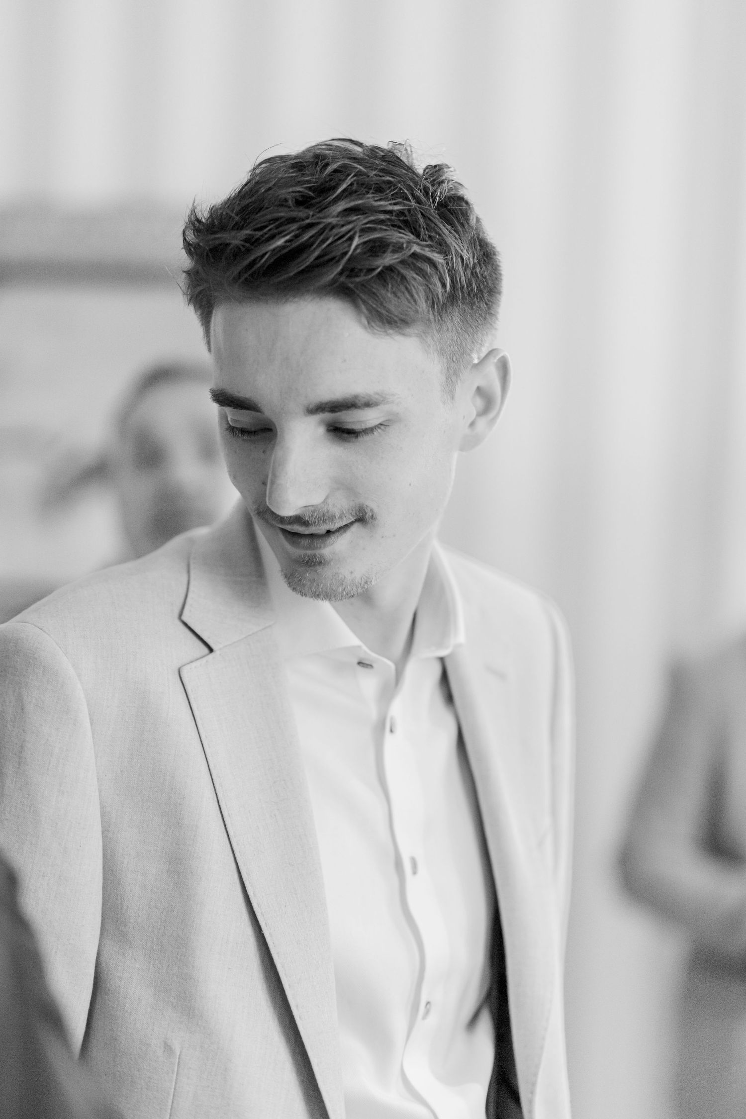 Black and white portrait of a person in a light colored suit jacket looking down with a subtle smile.