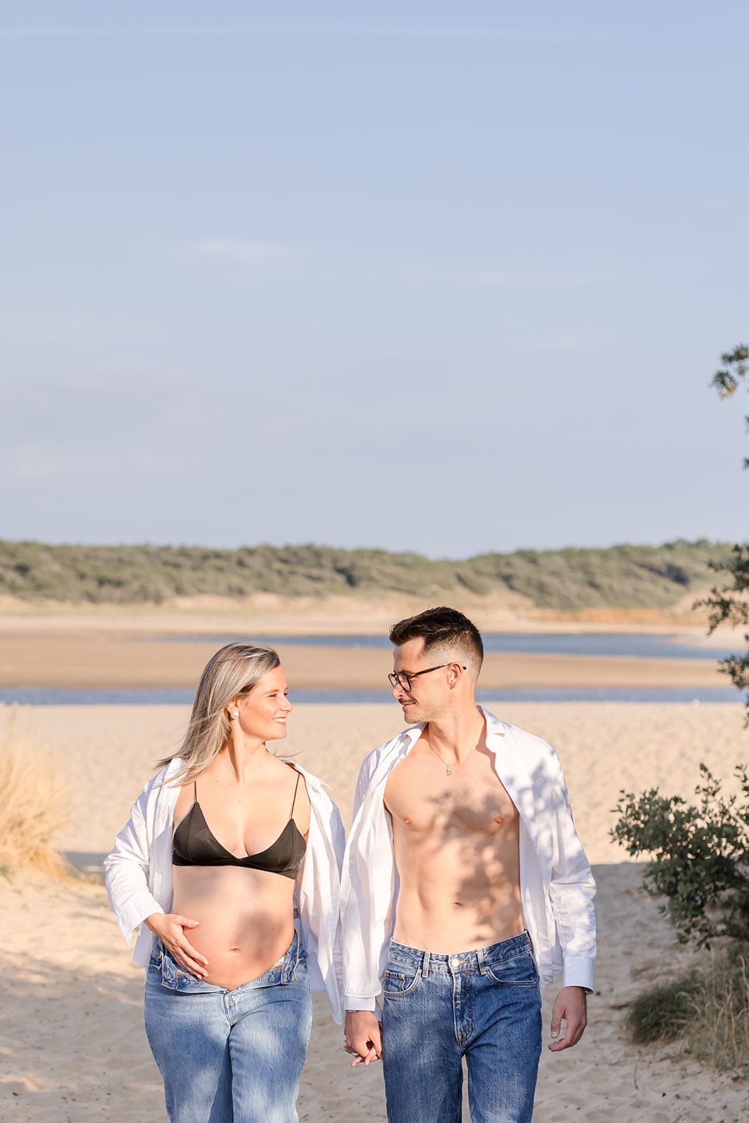 Couple walking and holding hands at a serene beach location with sand dunes in the background.