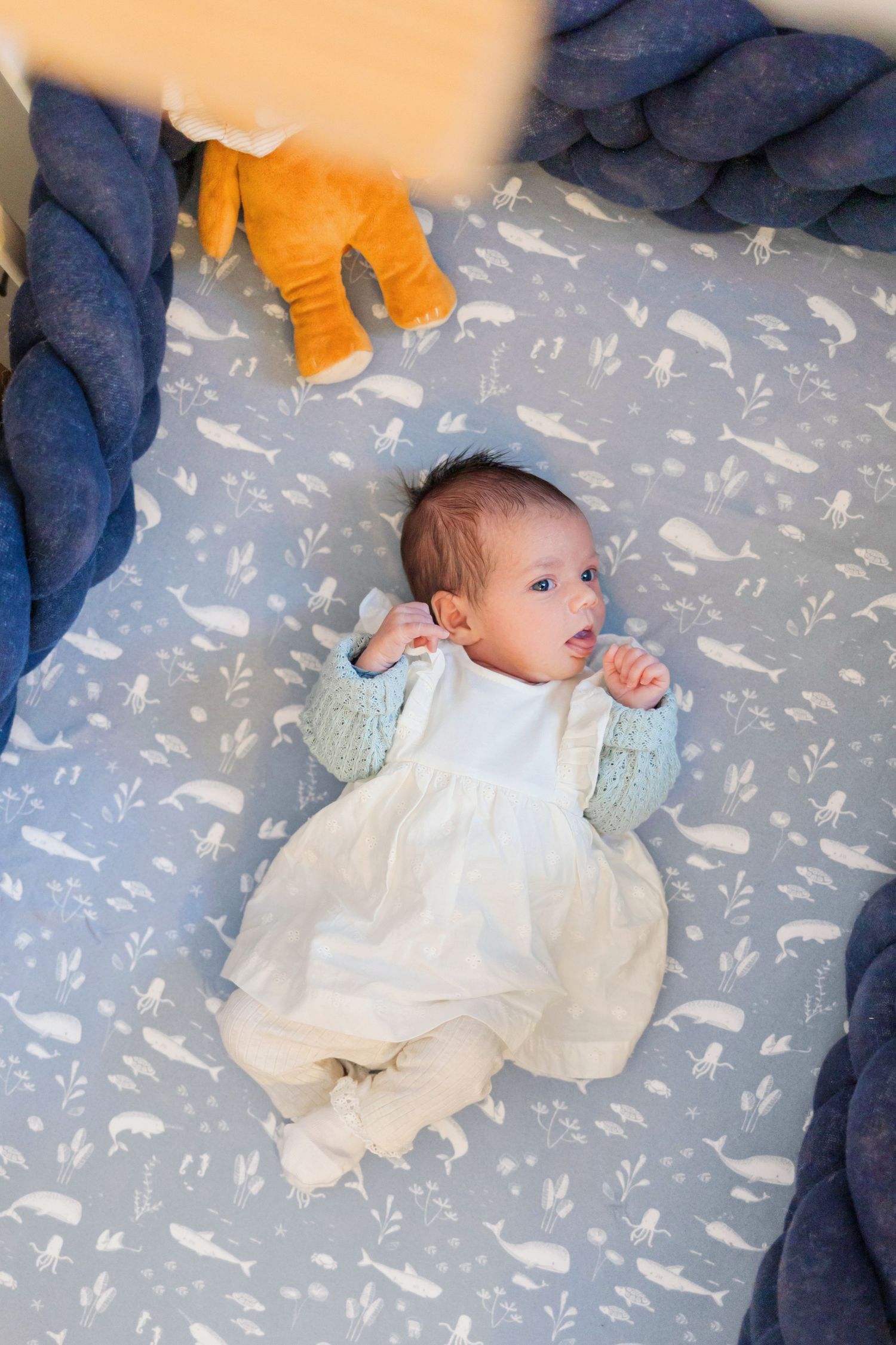 Infant wearing white dress and mint sweater lying on blue patterned bedding with orange plush toy nearby.
