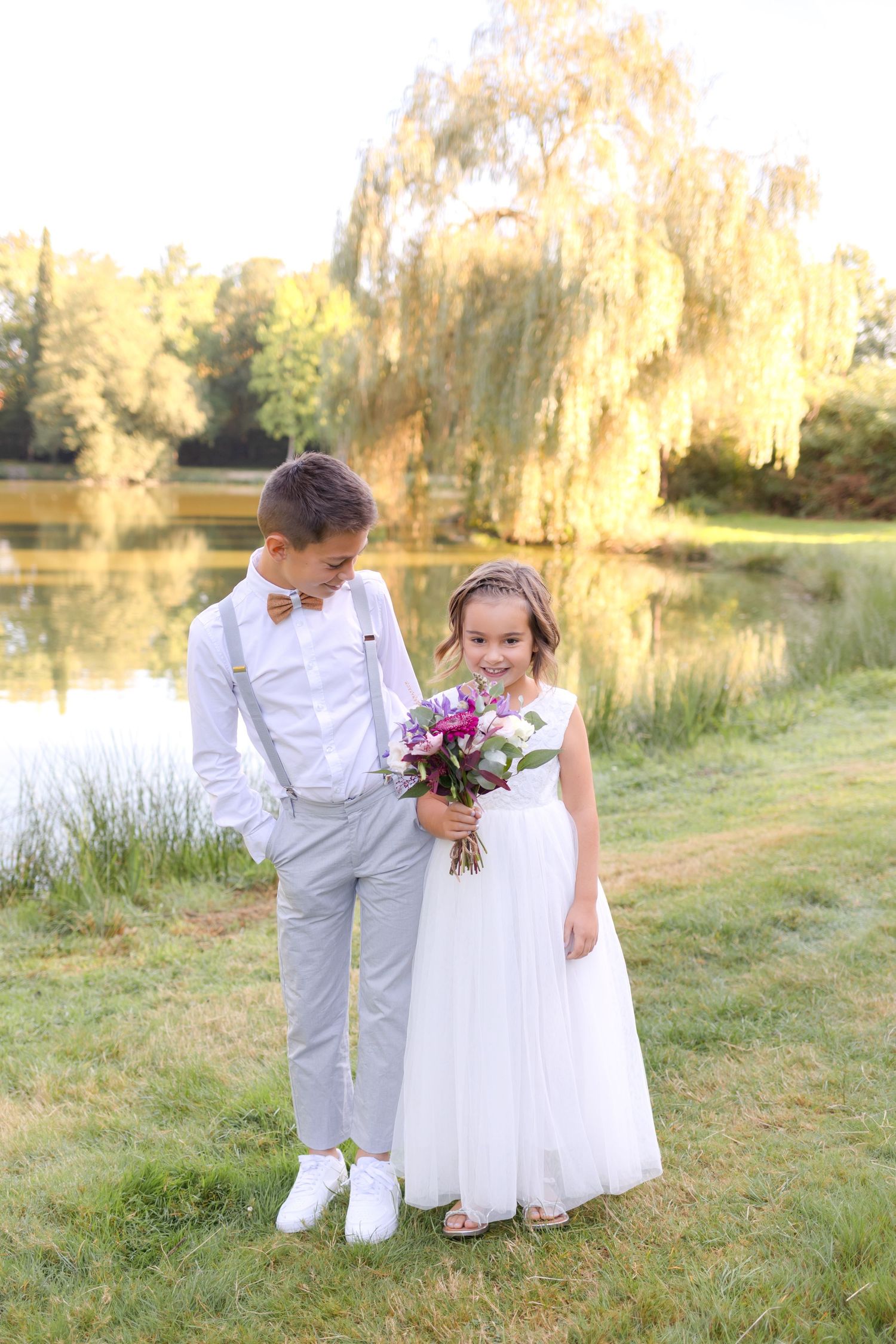 Two children in formal white attire stand by a pond with a willow tree in the background.