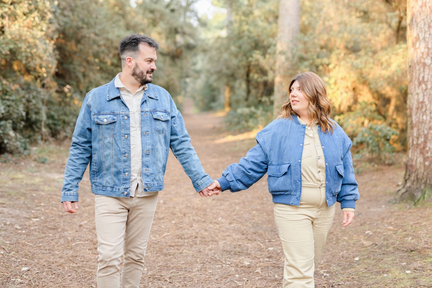 A couple wearing matching denim jackets walks hand in hand through a sunlit forest path.