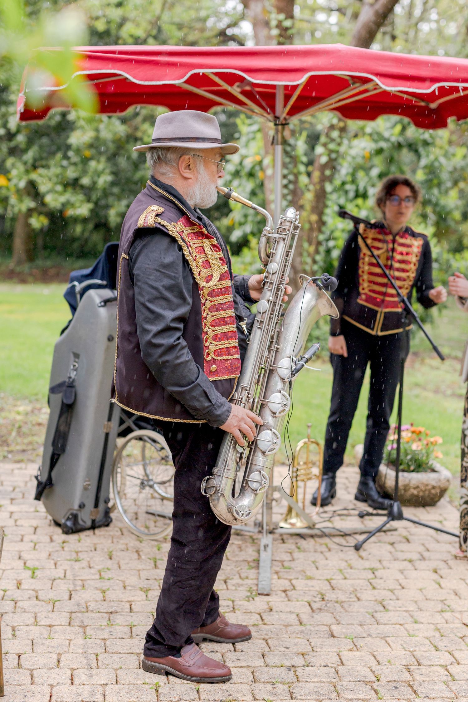Musicians in vintage uniforms with brass instruments performing under red umbrellas on cobblestone pavement.