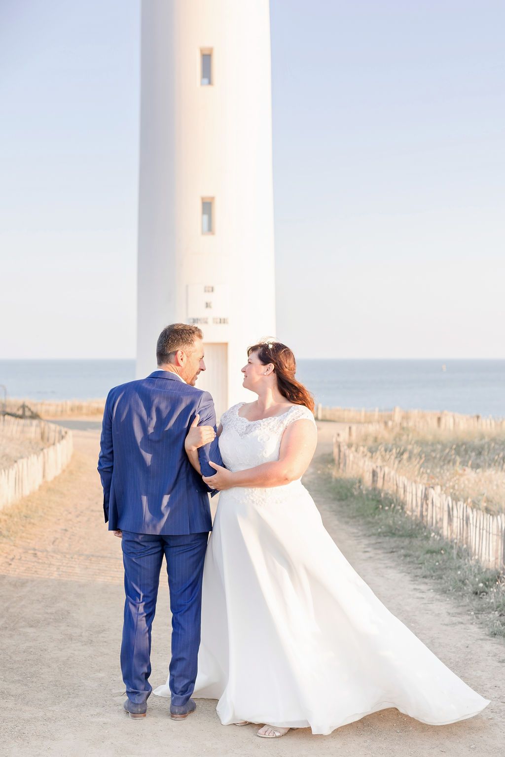Newlyweds share a romantic moment on a path next to a coastal lighthouse.