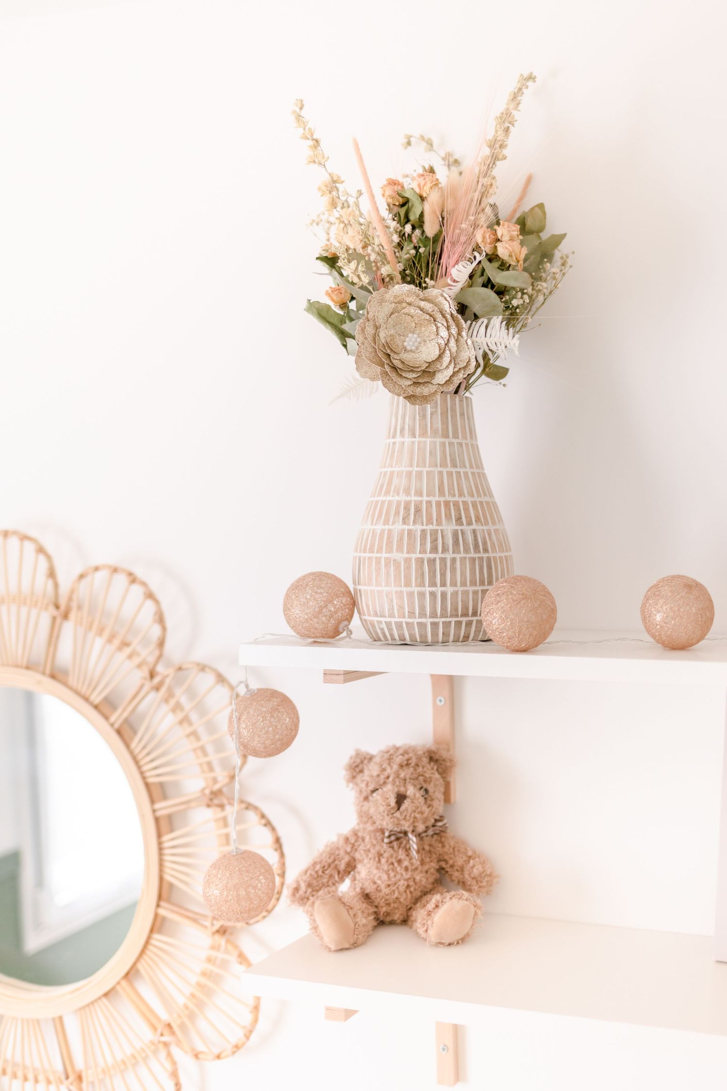 Textured vase with dried flowers on white shelf with rattan mirror and teddy bear decor.