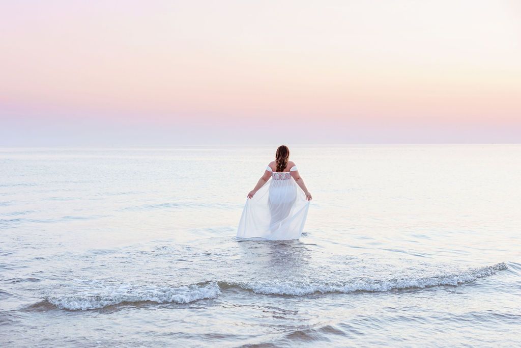Woman in flowing white dress walks deeper into ocean waves during sunset photo session.