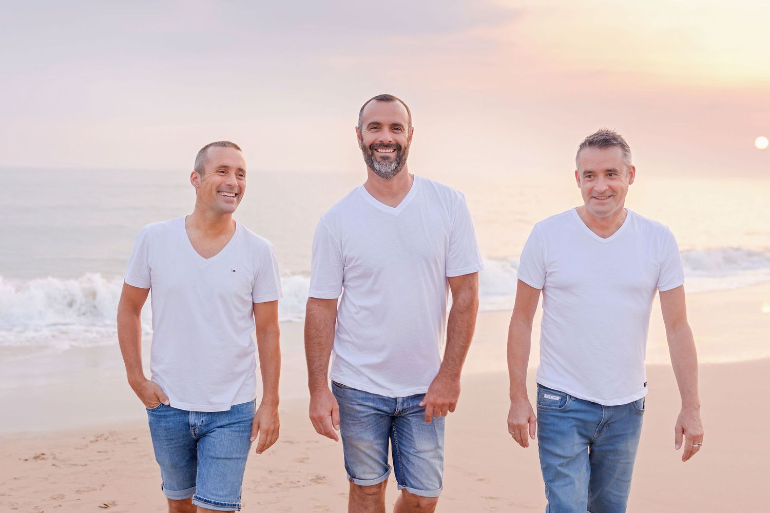 Three people in matching white shirts and denim walking on beach during golden hour.