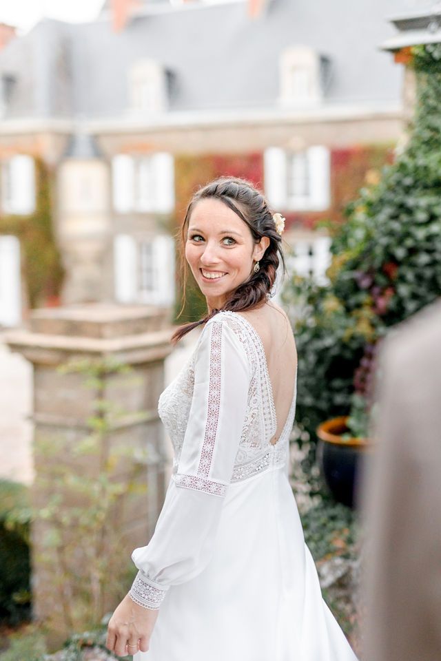 Bride in lace-detailed wedding dress smiling over shoulder in garden setting.
