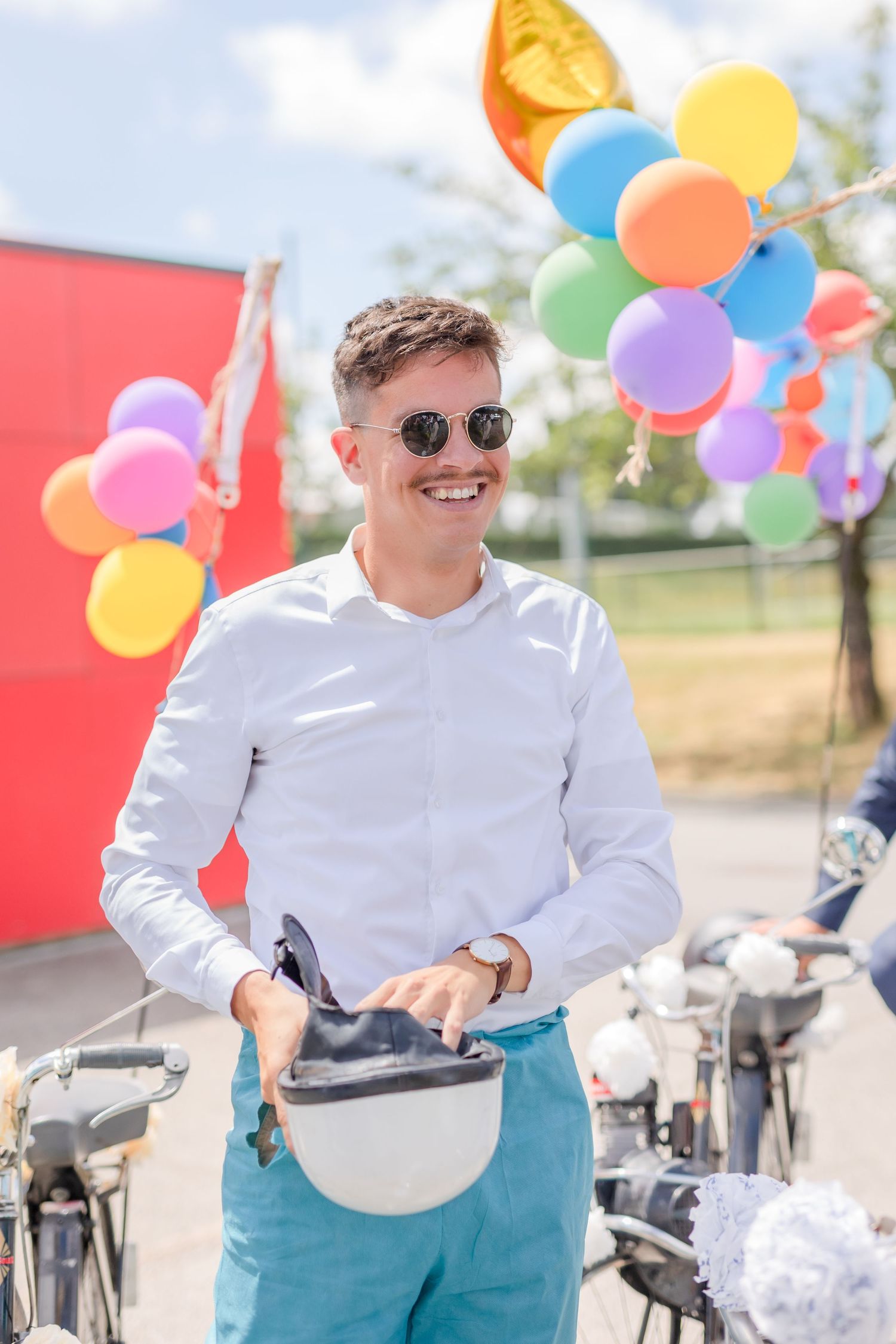 Guest in white shirt and turquoise pants poses with balloons near bicycles at outdoor event.