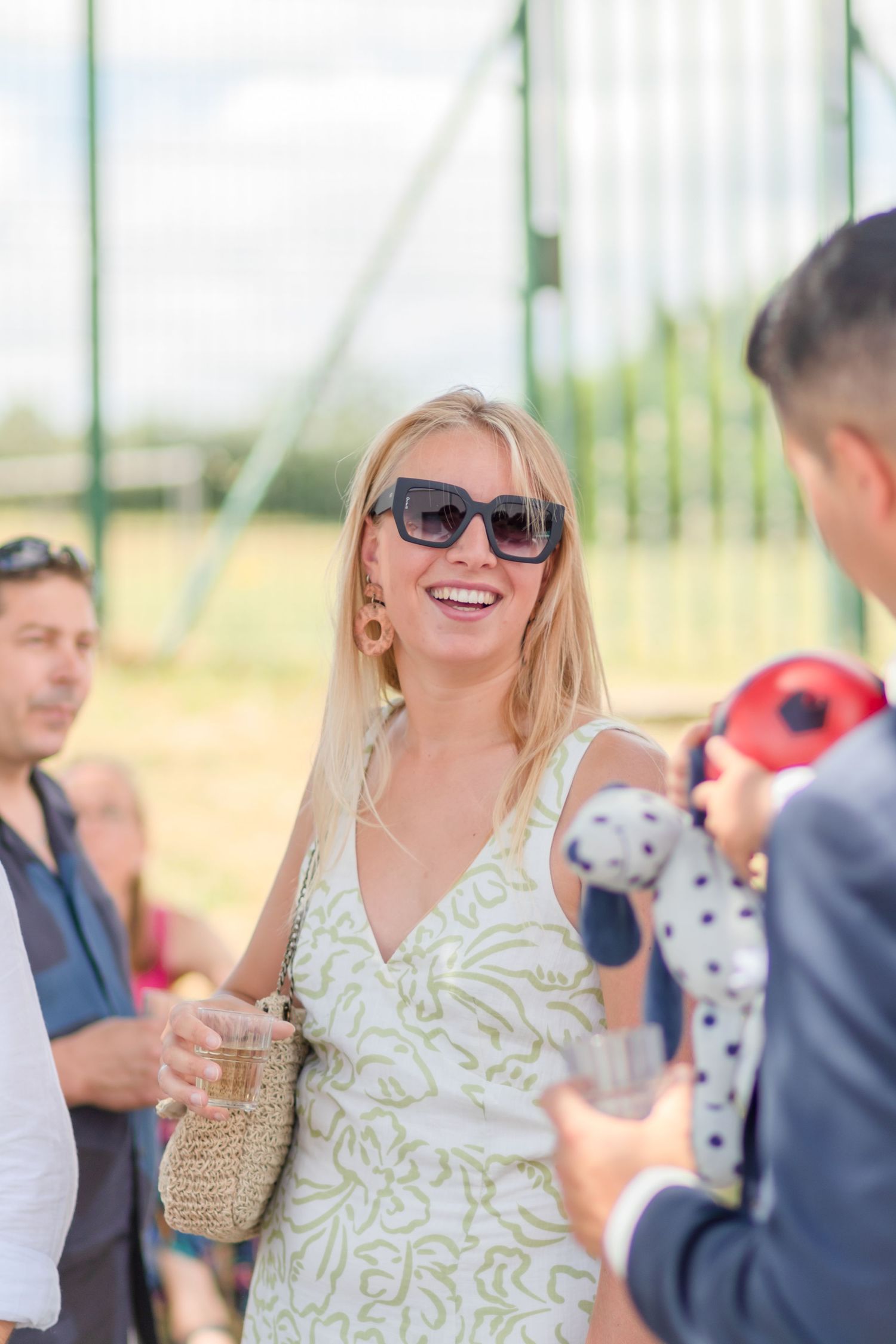 Outdoor summer party guest in white dress and sunglasses holding woven bag.
