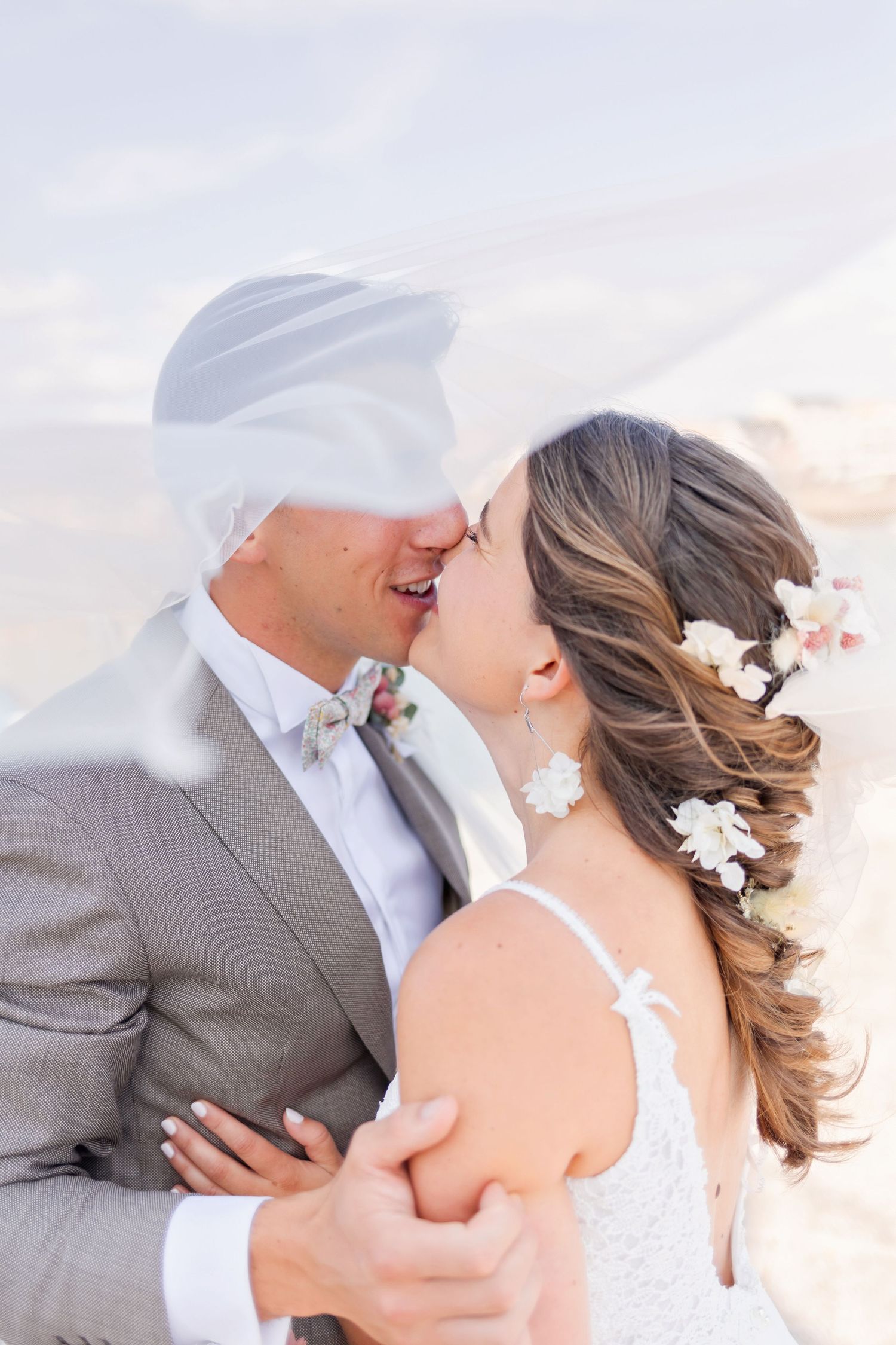 Intimate wedding moment captured as couple embraces under flowing veil by the sea.
