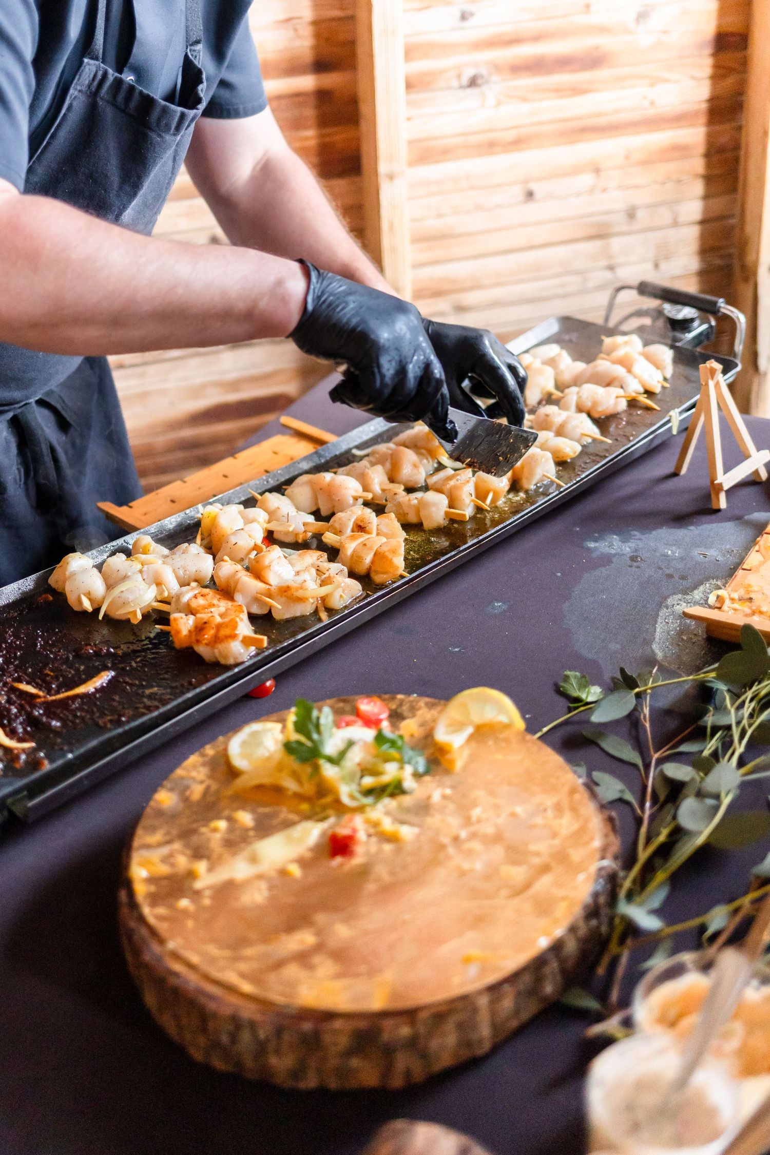 Chef in black gloves preparing food on a long serving tray next to a wooden platter.