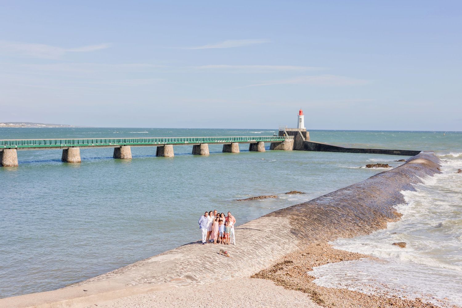 Long pier stretches into blue ocean with lighthouse at end and small figures on beach.