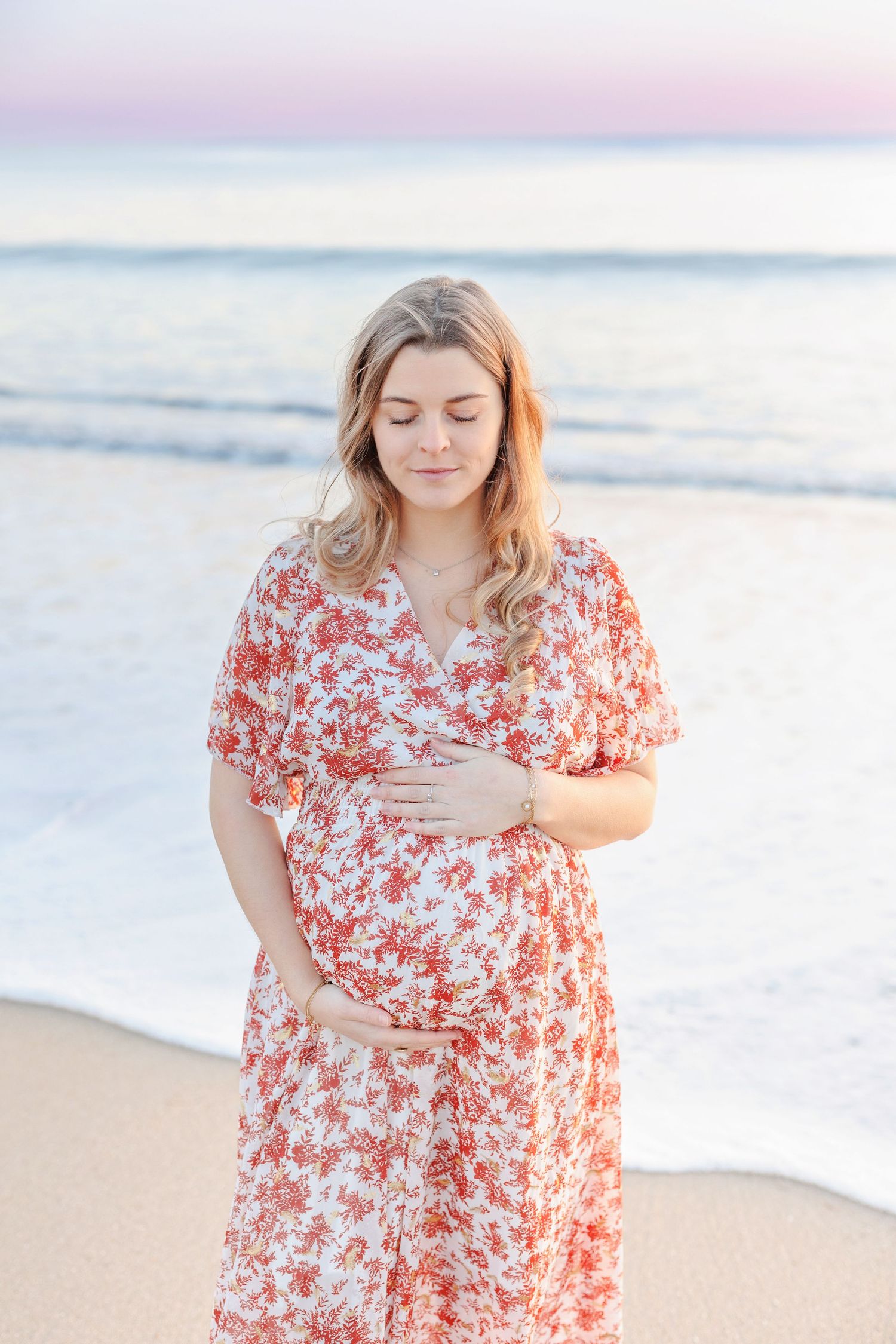 A person in a flowing floral dress stands on the beach during golden hour.