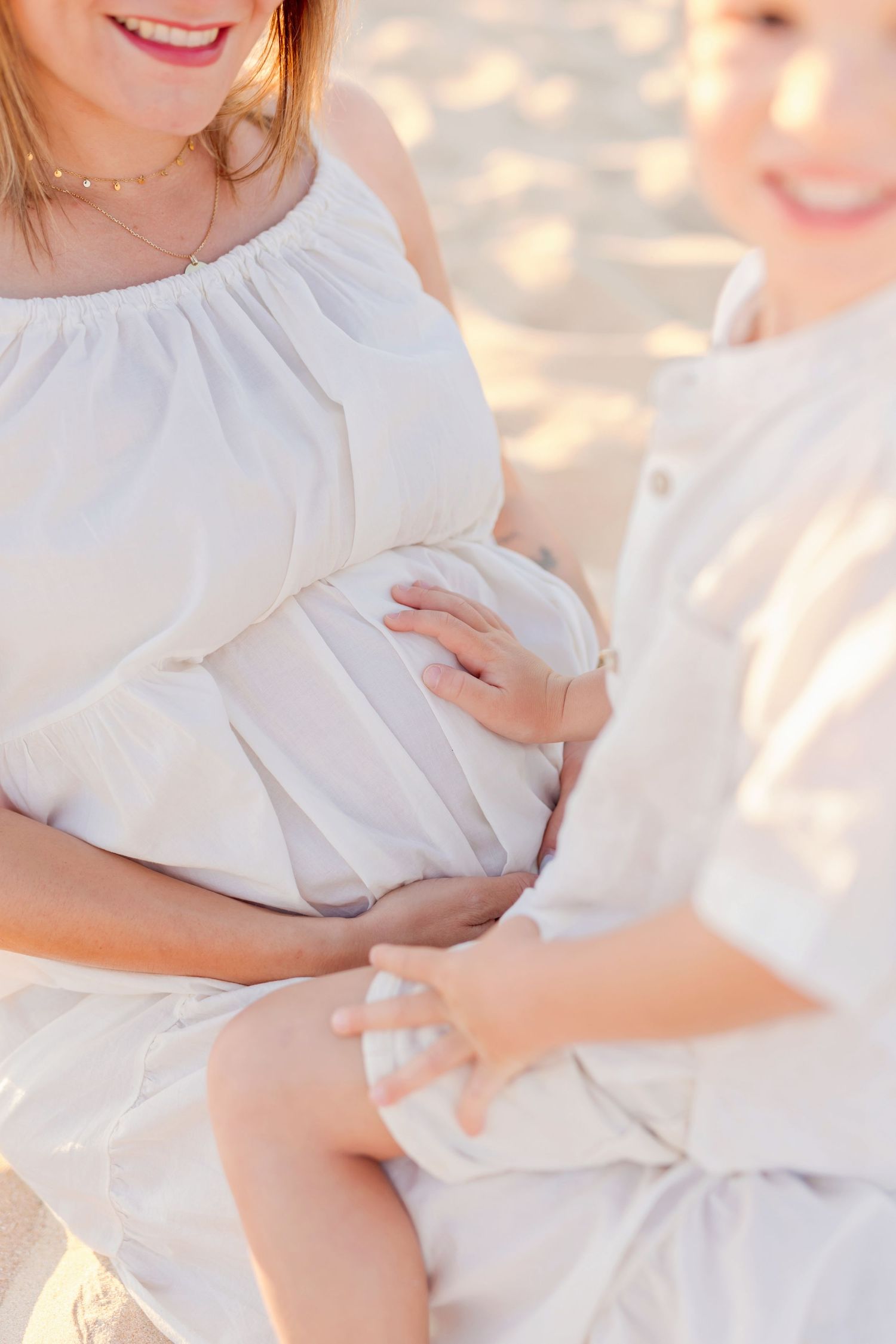 Close-up of pregnancy moment captured in white dress on sunny beach.