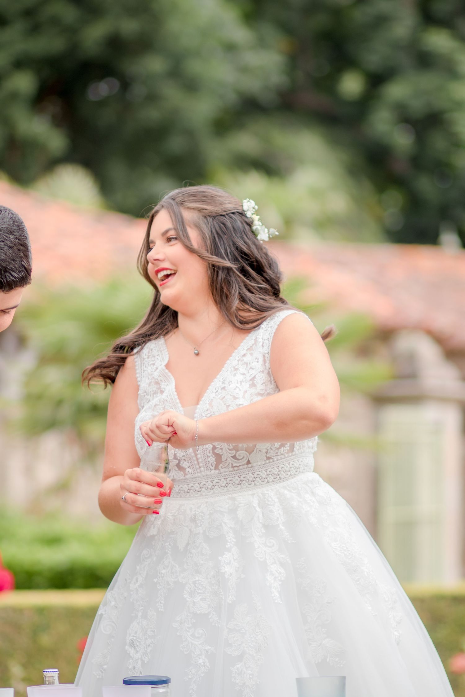 A bride in a white lace dress laughs joyfully during an outdoor wedding celebration.