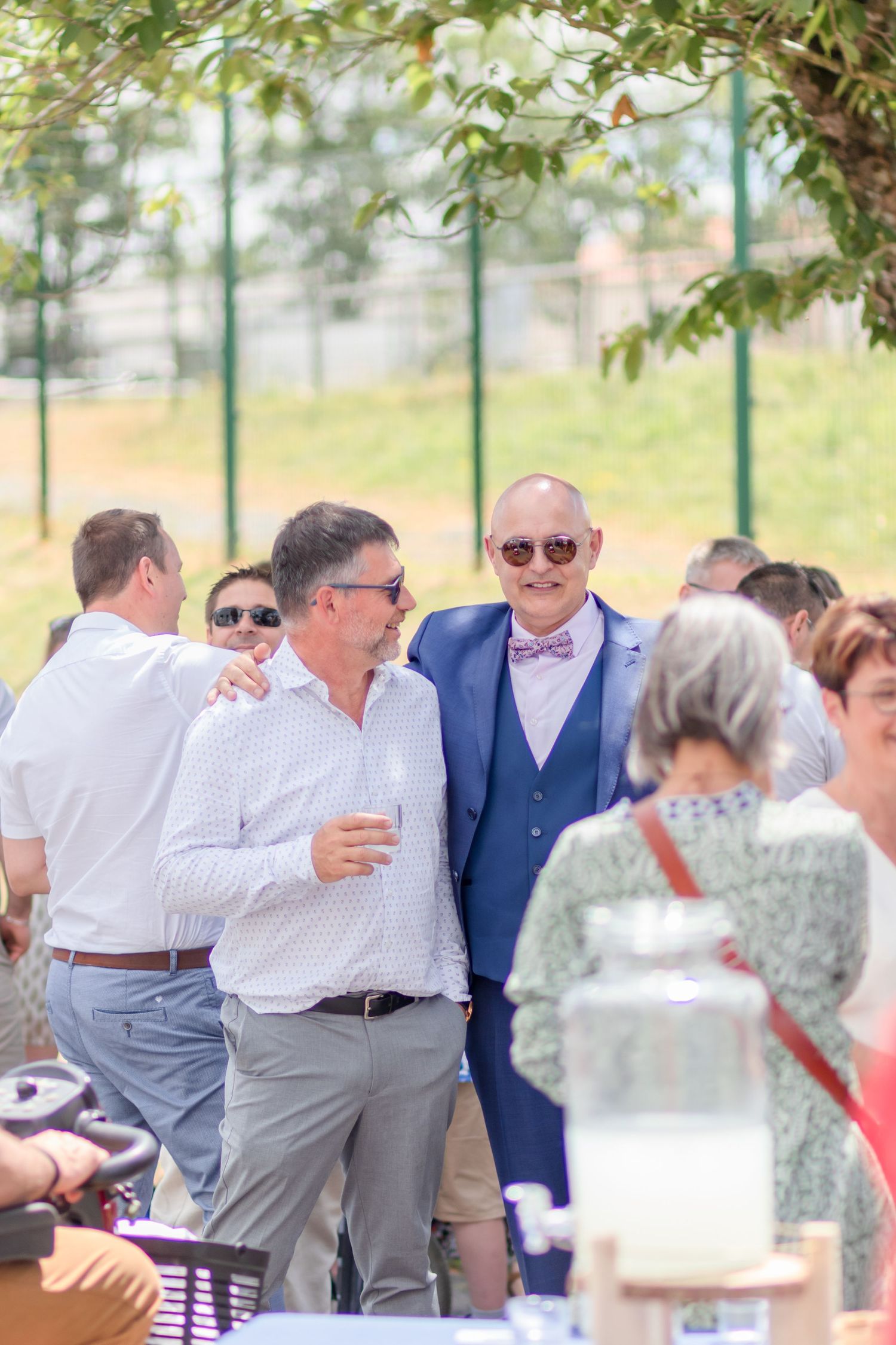 Wedding guests socializing at outdoor reception with greenery and fence in background.