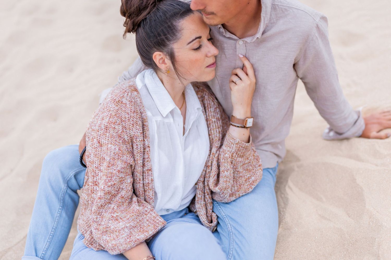 Intimate beach scene with couple in casual attire sharing tender moment against sandy backdrop.