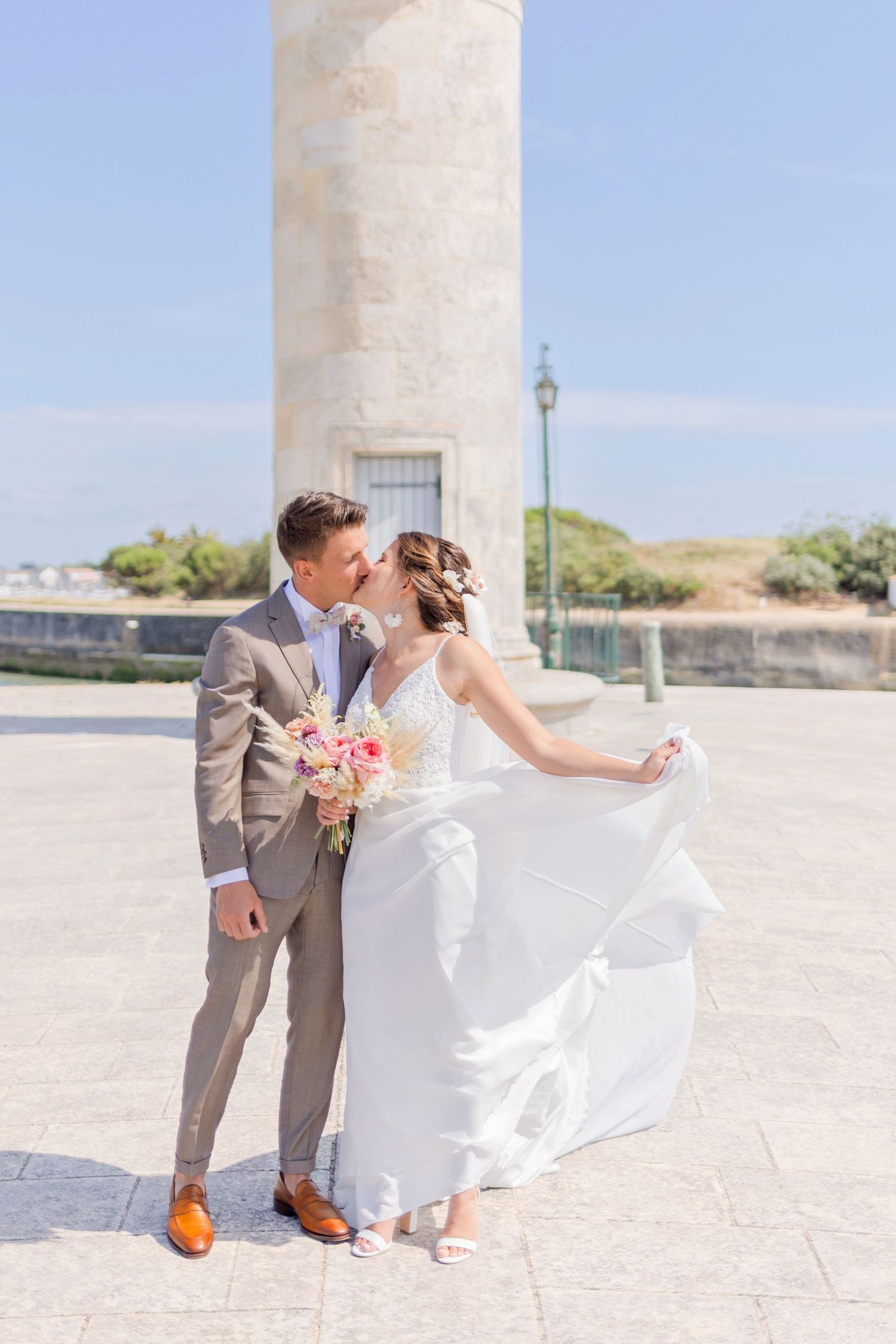 Wedding couple poses by historic lighthouse in elegant attire as bride's dress flows in wind.