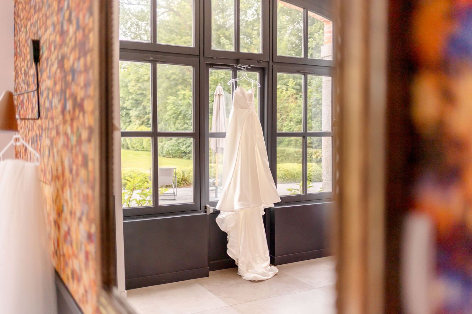 White wedding dress hanging in sunlit window with floral wallpaper backdrop.