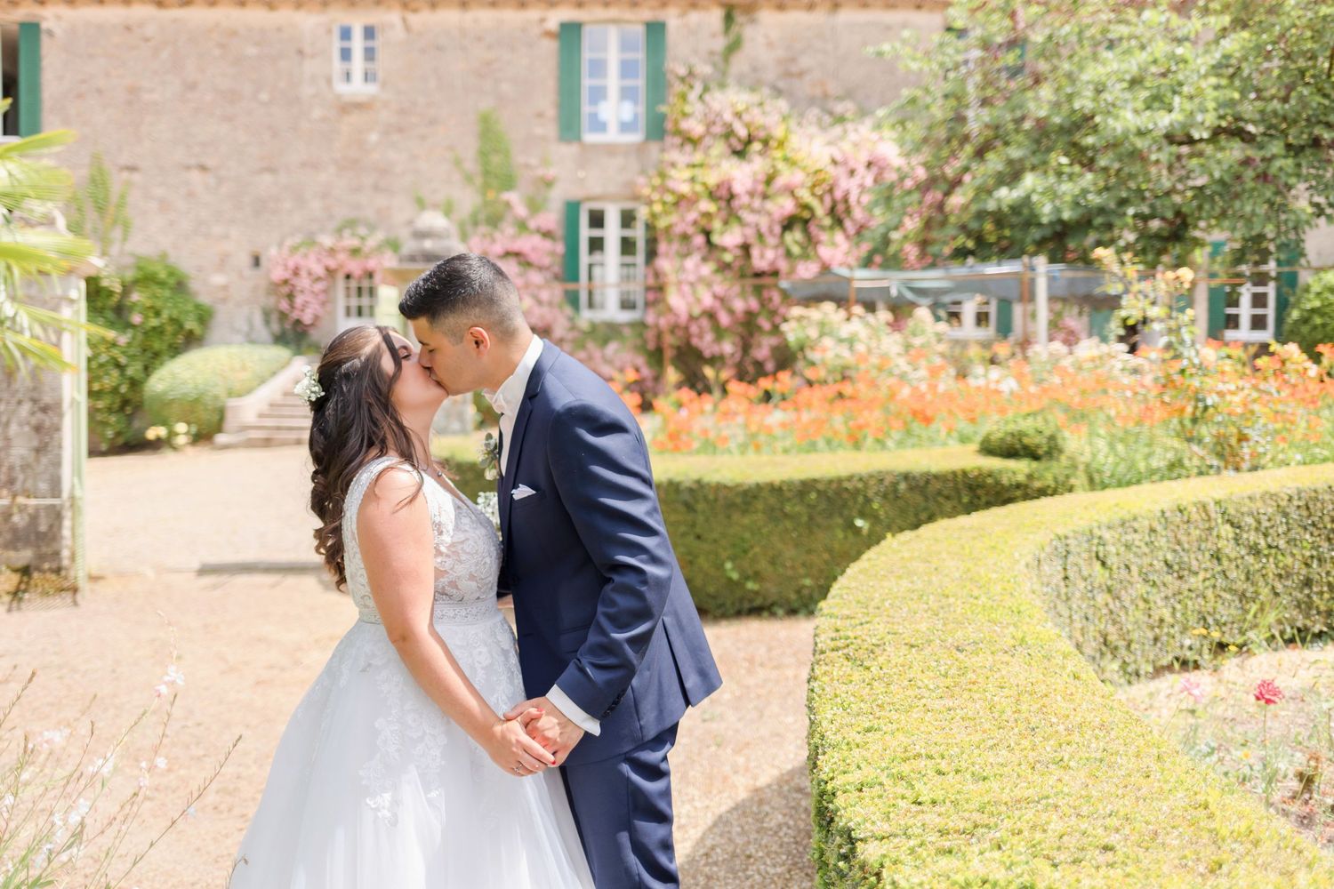 A romantic wedding moment captured in a lush garden setting with formal hedges and flowering plants.