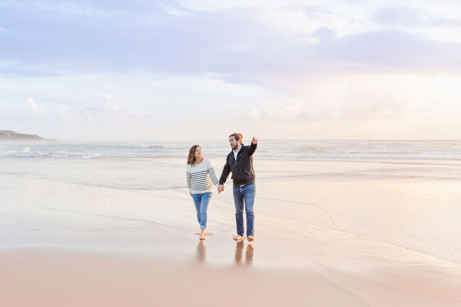Couple walking along shoreline during sunset with waves crashing in background.
