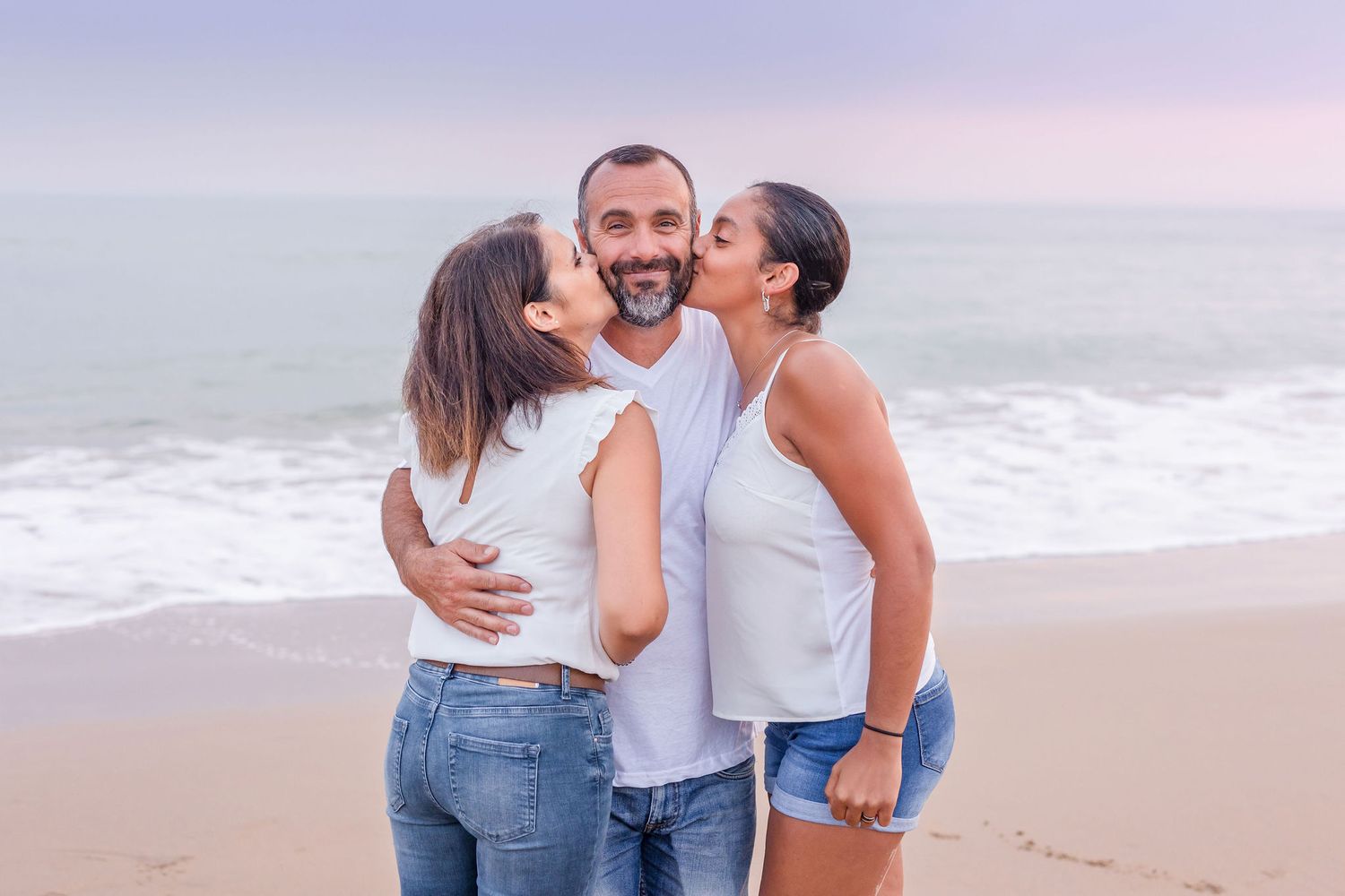 Three people sharing an affectionate moment on beach at sunset in casual attire.